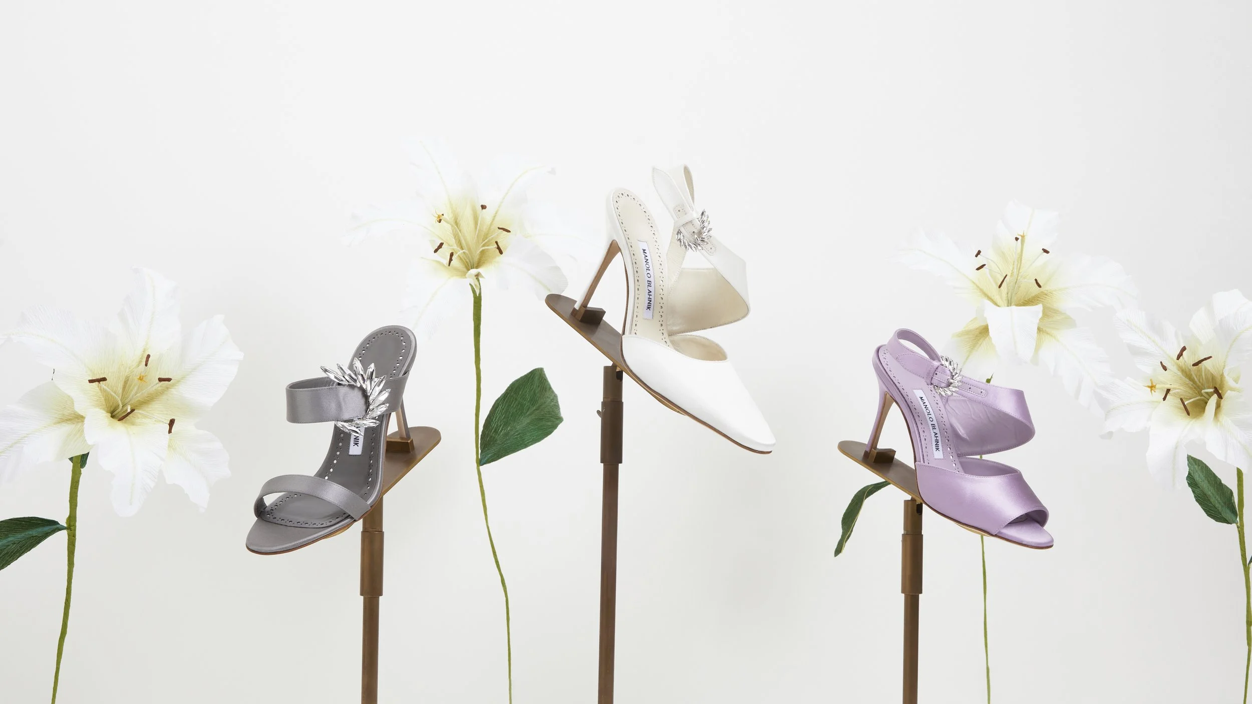 Display of three high-heeled shoes with floral accents on stands, surrounded by white paper flowers with green stems and leaves by Emmeli Kimhi