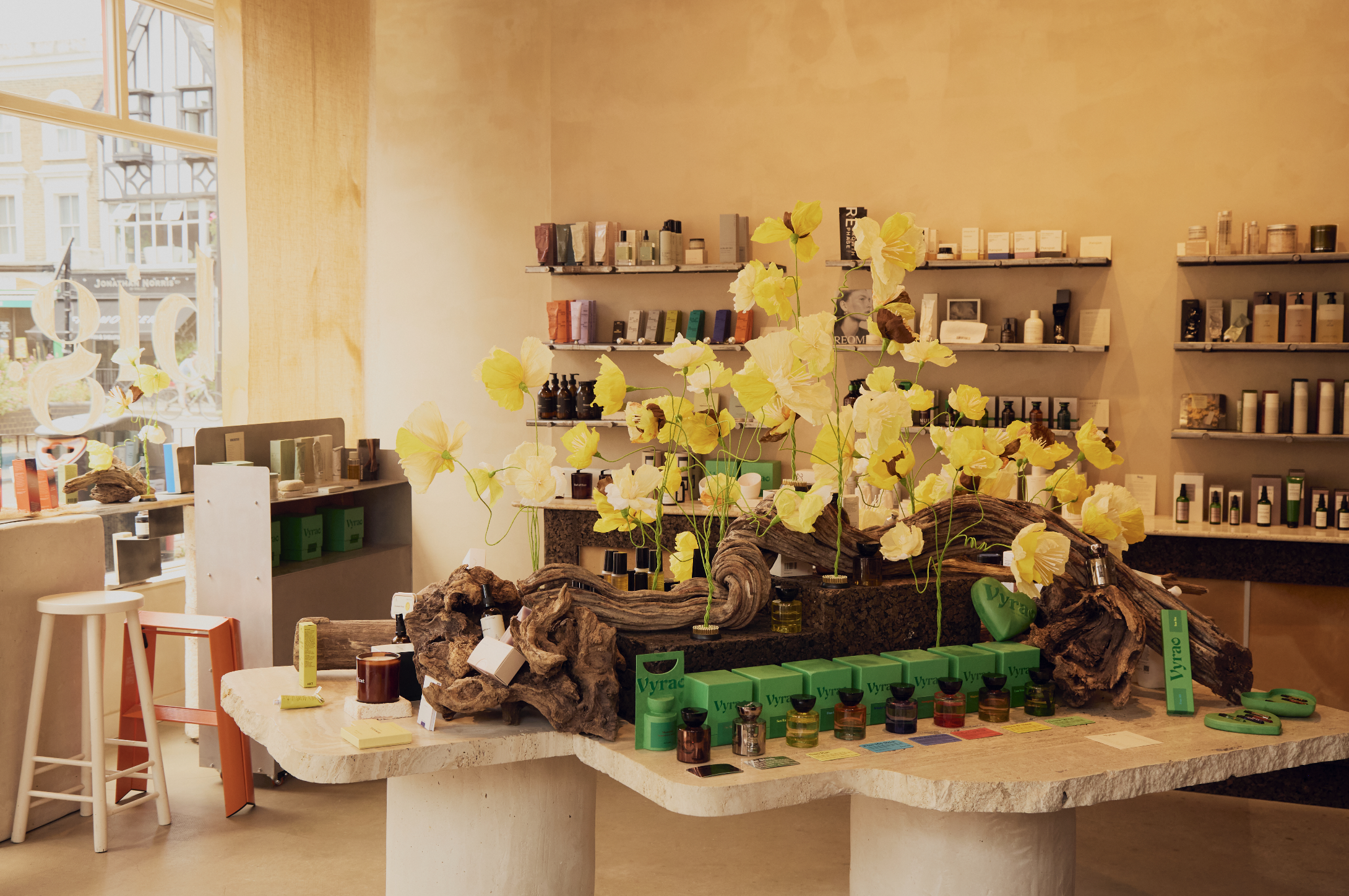 Handmade yellow paper poppy installation by Emmeli Kimhi displayed on a large driftwood sculpture atop a marble table inside Big In Hackney store in London, surrounded by product shelves and packaging