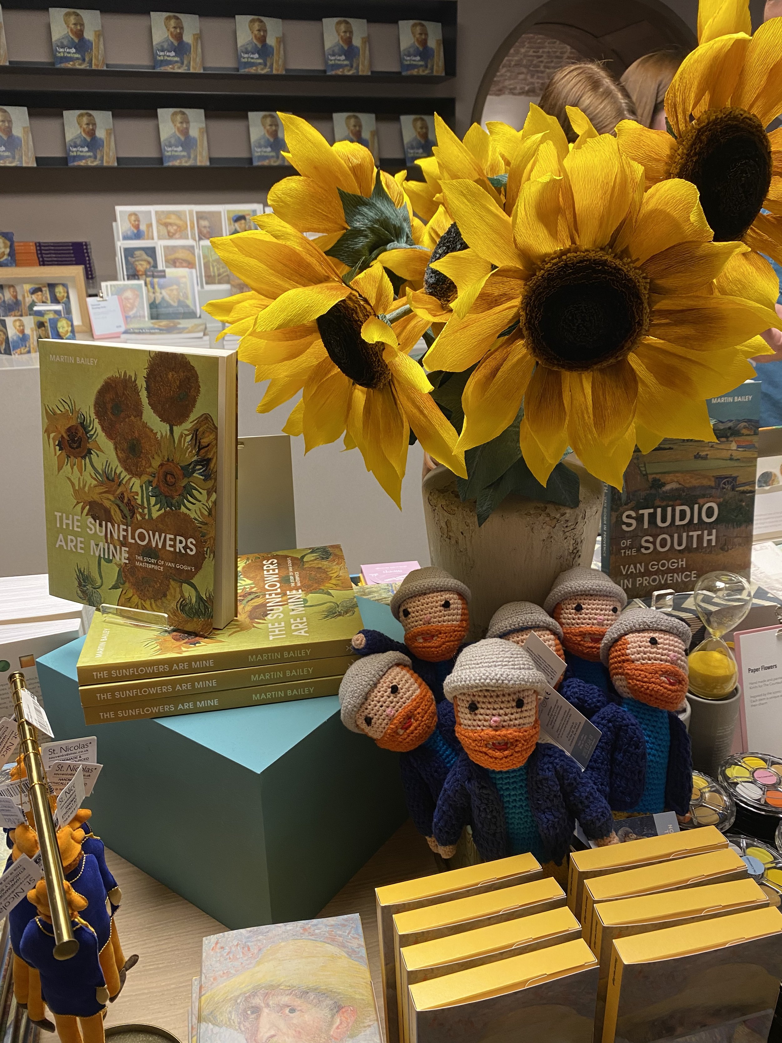 A display table with books about Vincent van Gogh, including "The Sunflowers Are Mine." There is a large bouquet of paper sunflowers by Emmeli Kimhi and crocheted doll figures. In the background, shelves hold books and images of Van Gogh.
