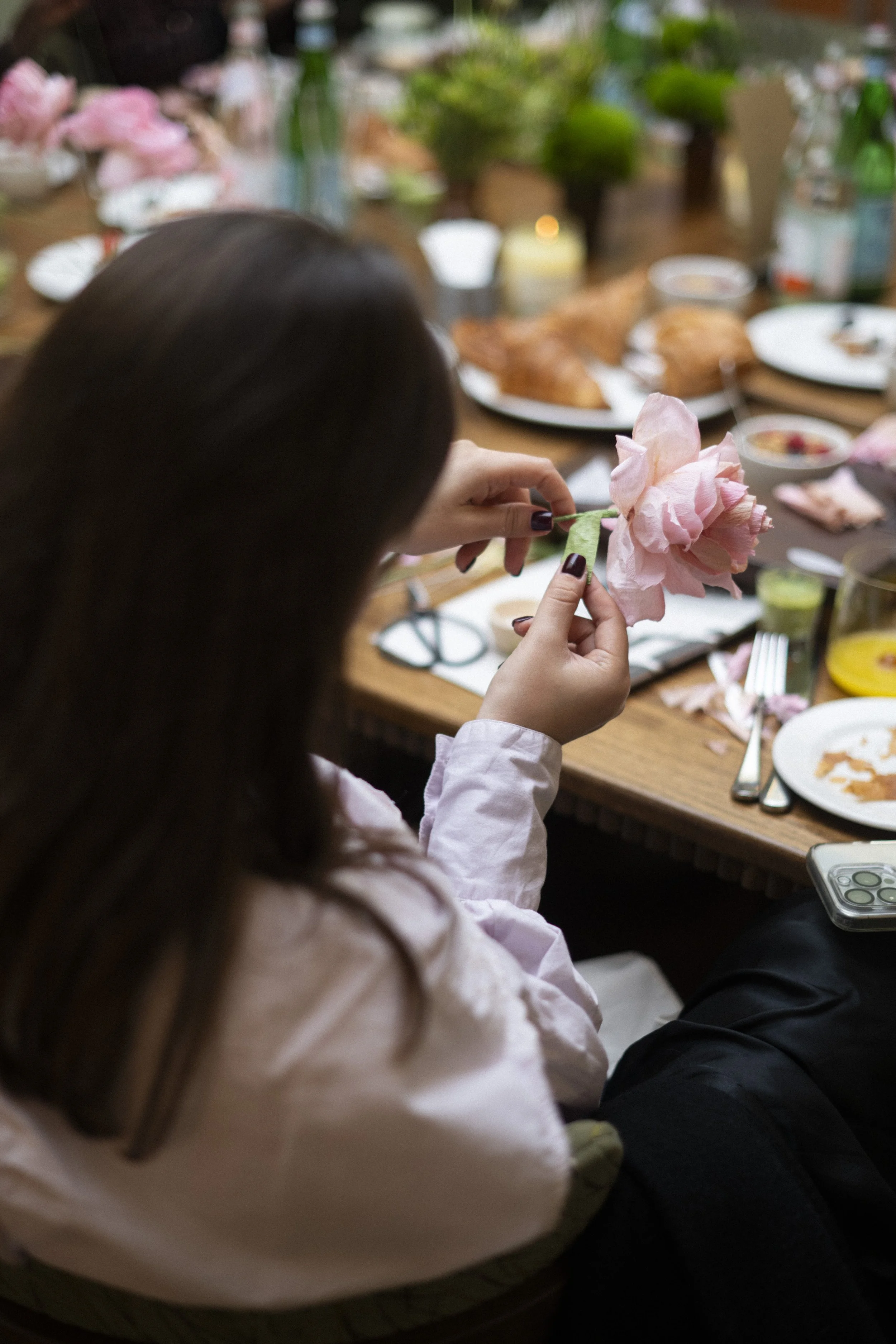 Person holding a pink flower, setting on a table with dishes and food, surrounded by a dining setup. Emmeli Kimhi's Le Labo workshop.