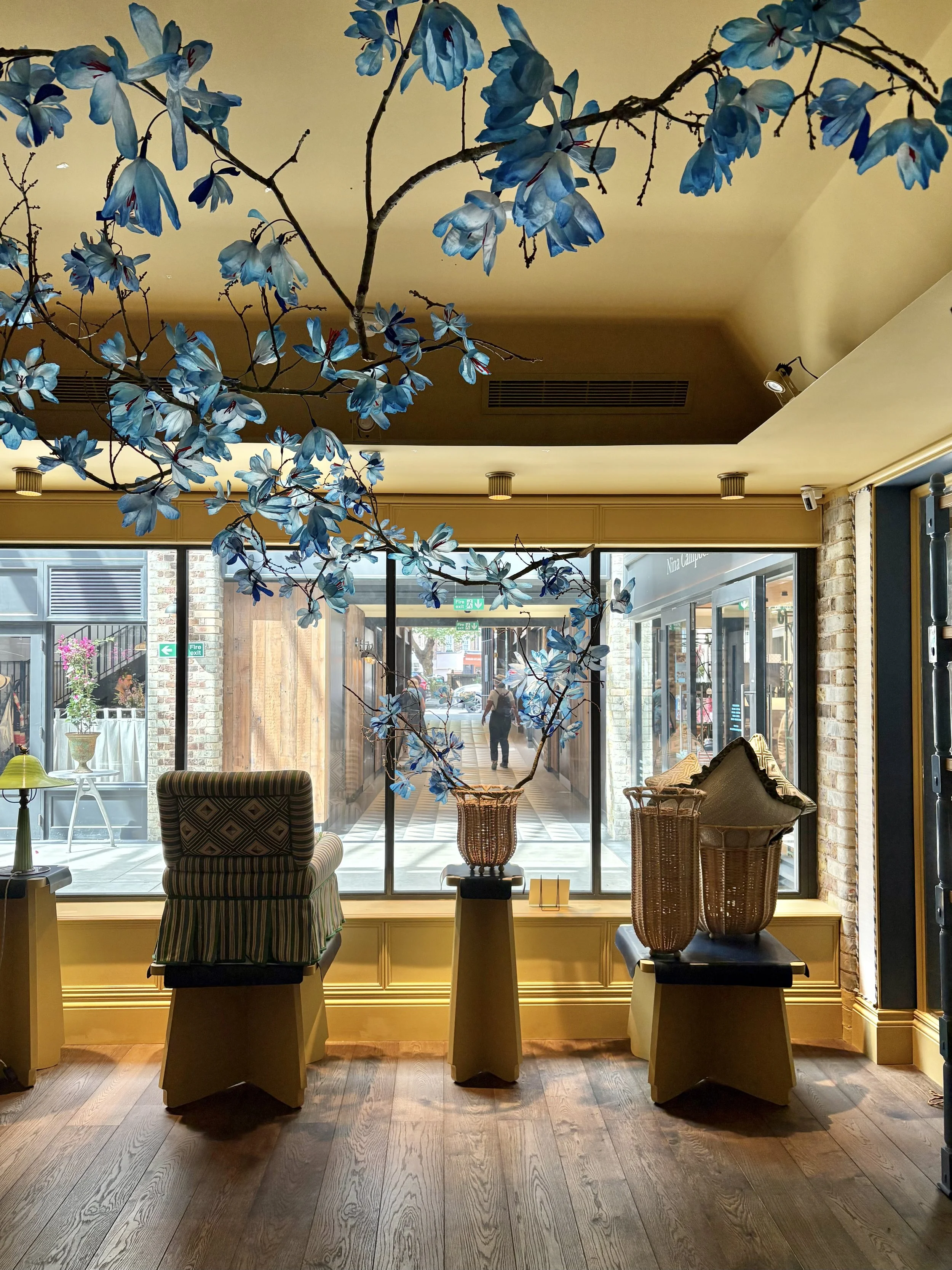 Large handmade blue paper magnolia branch installation by Emmeli Kimhi hanging from the ceiling in a bright retail space at And Objects, London, with striped armchair and wicker baskets below
