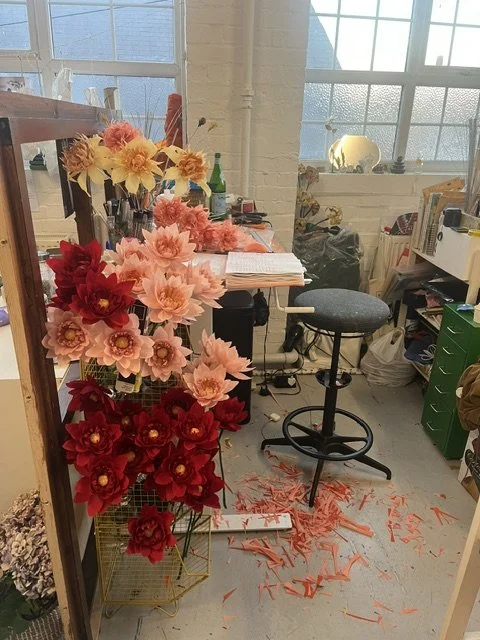 Emmeli Kimhi's workspace with a colorful display of artificial flowers on a metal stand, with shredded orange paper on the floor, a high adjustable stool, and various supplies and books in a bright room with large windows.