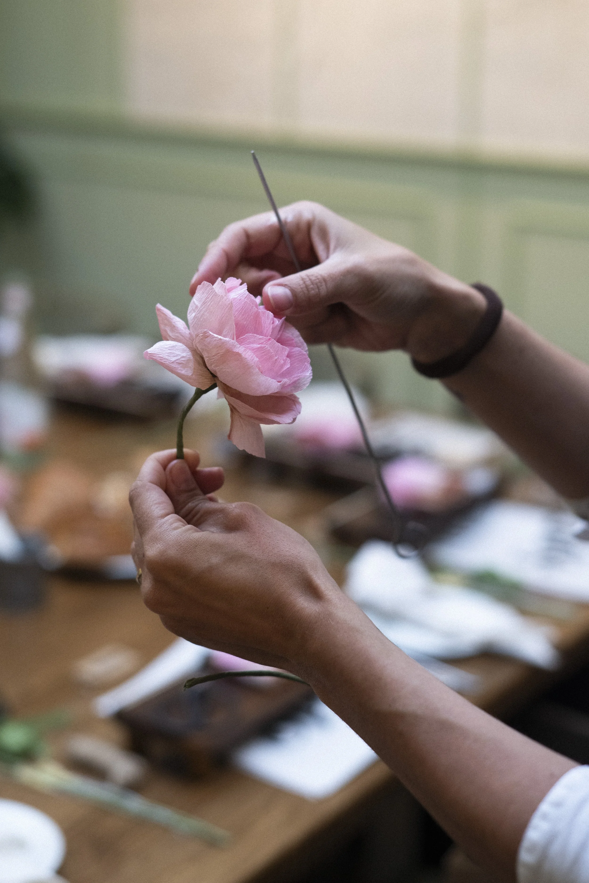 Emmeli Kimhi is carefully designing a pink paper flower, likely a peony, with a needle on a crafting table filled with supplies.