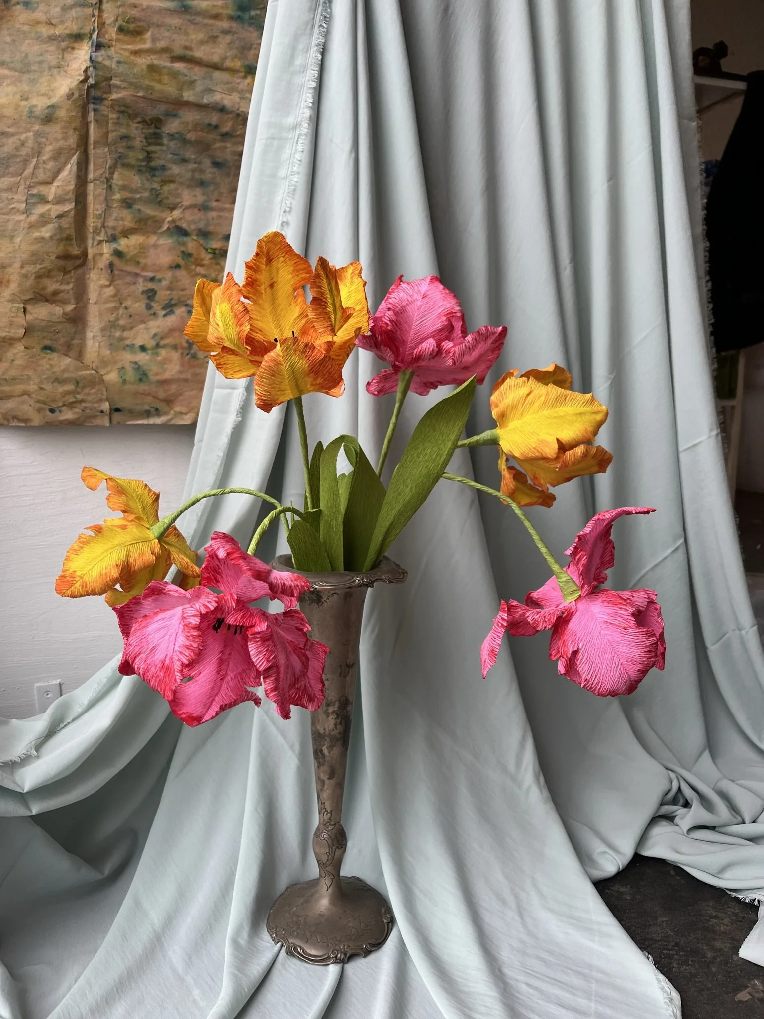A bouquet of orange, pink, and yellow flowers by Emmeli Kimhi in an ornate silver vase placed on light-colored draped fabric, with a brown abstract painting hanging on the wall behind.