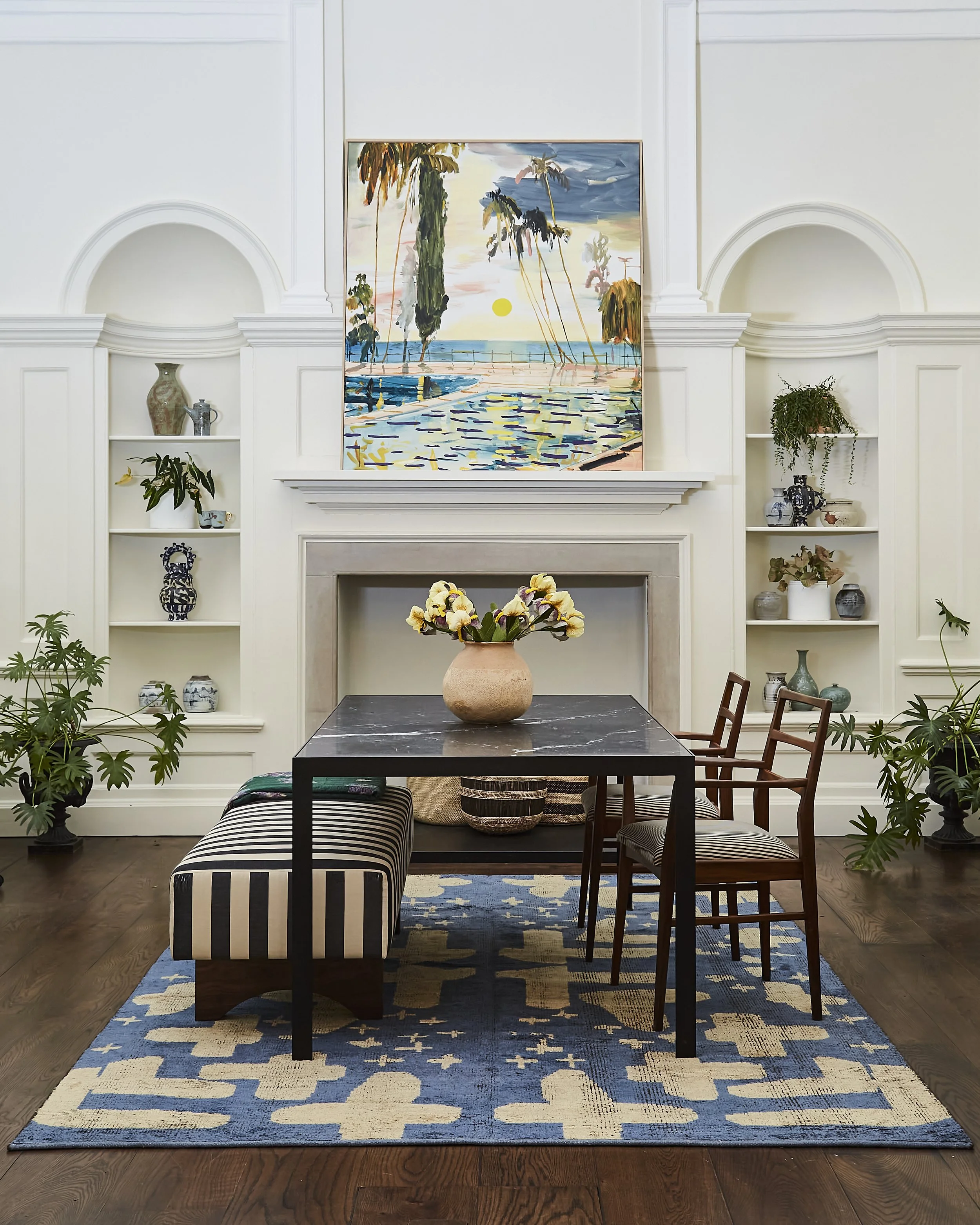 A stylish living room with a large blue and cream geometric patterned area rug, a dining table with a flower vase, wooden chairs, and a fireplace with built-in shelves holding various decorative items. Above the fireplace is a large colorful painting