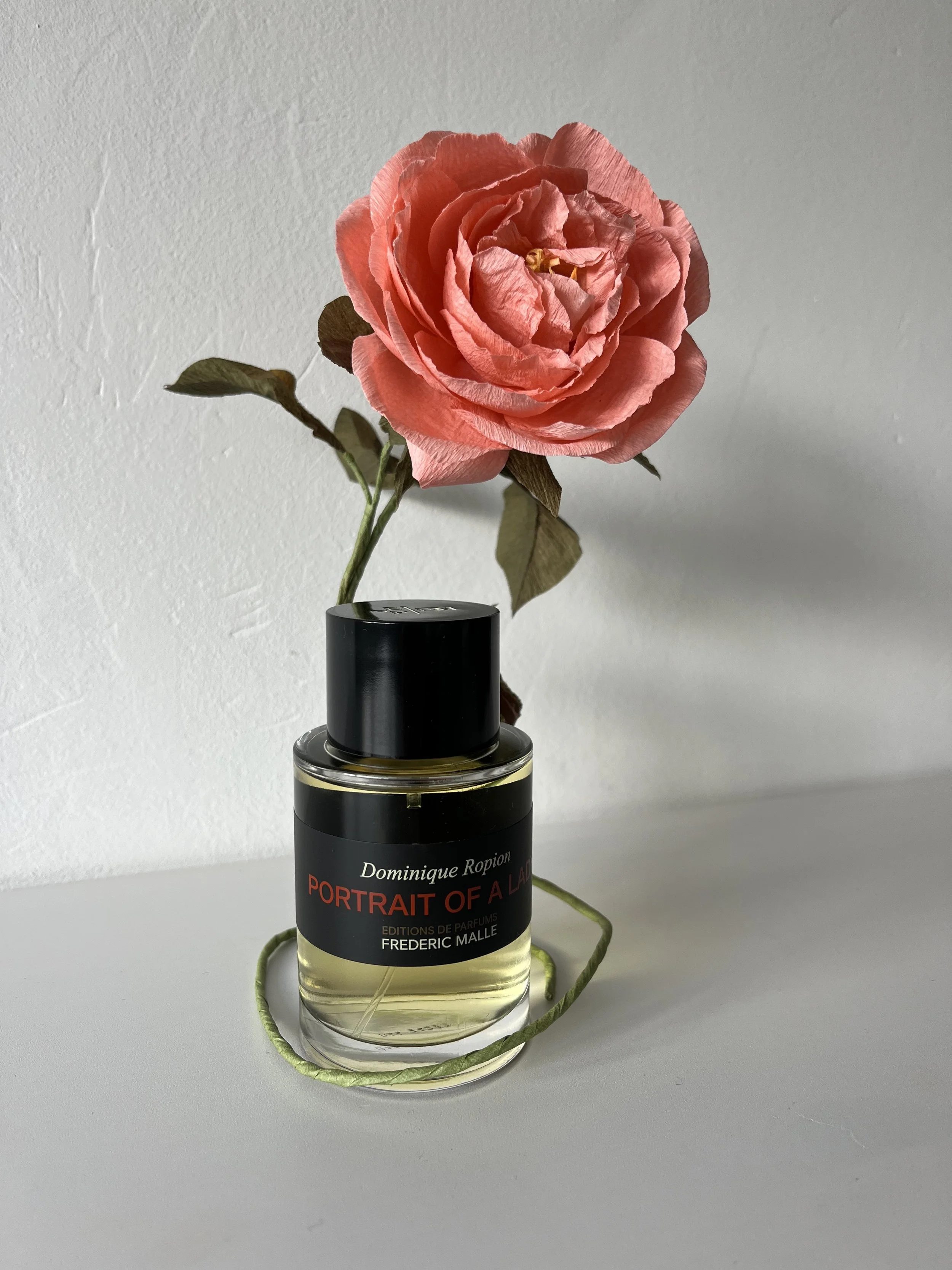 A perfume bottle labeled 'Portrait of a Lady' by Frederic Malle with a pink artificial rose flower  by Emmeli Kimhi and green decorative string on a white surface in front of a white wall.