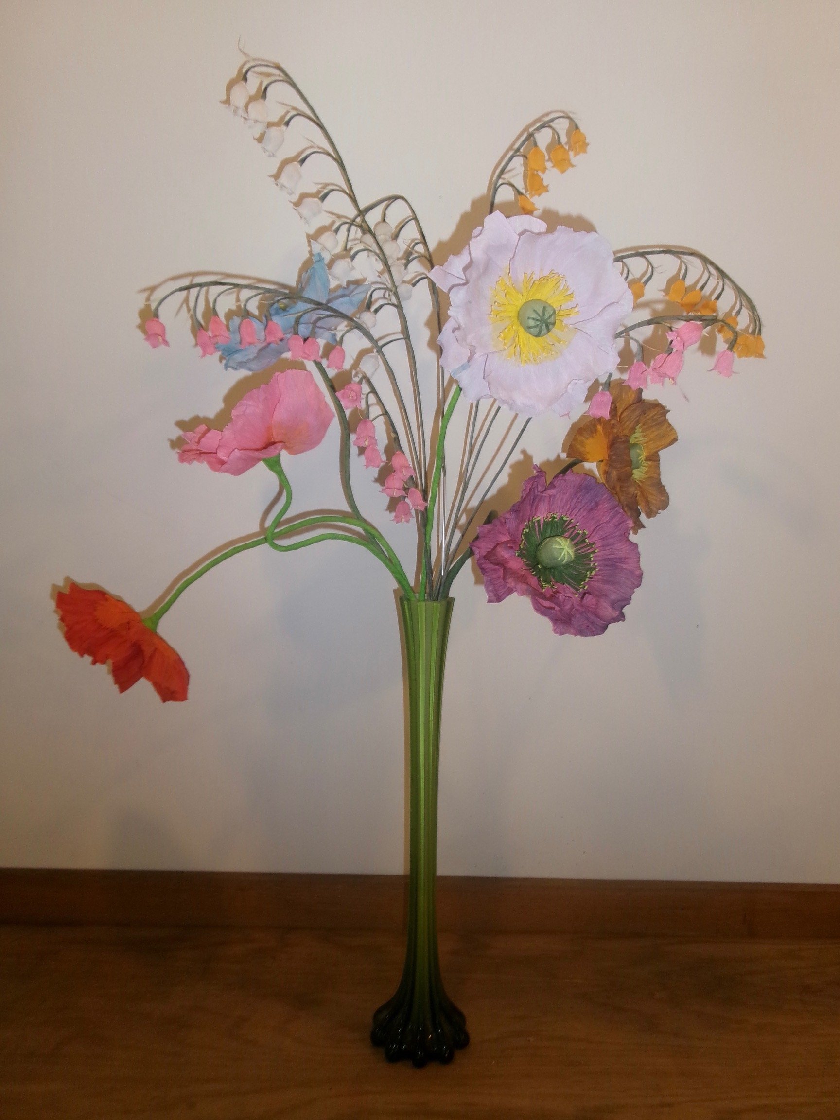 Decorative paper flower arrangement by Emmeli Kimhi in a green vase on a wooden surface with a plain background
