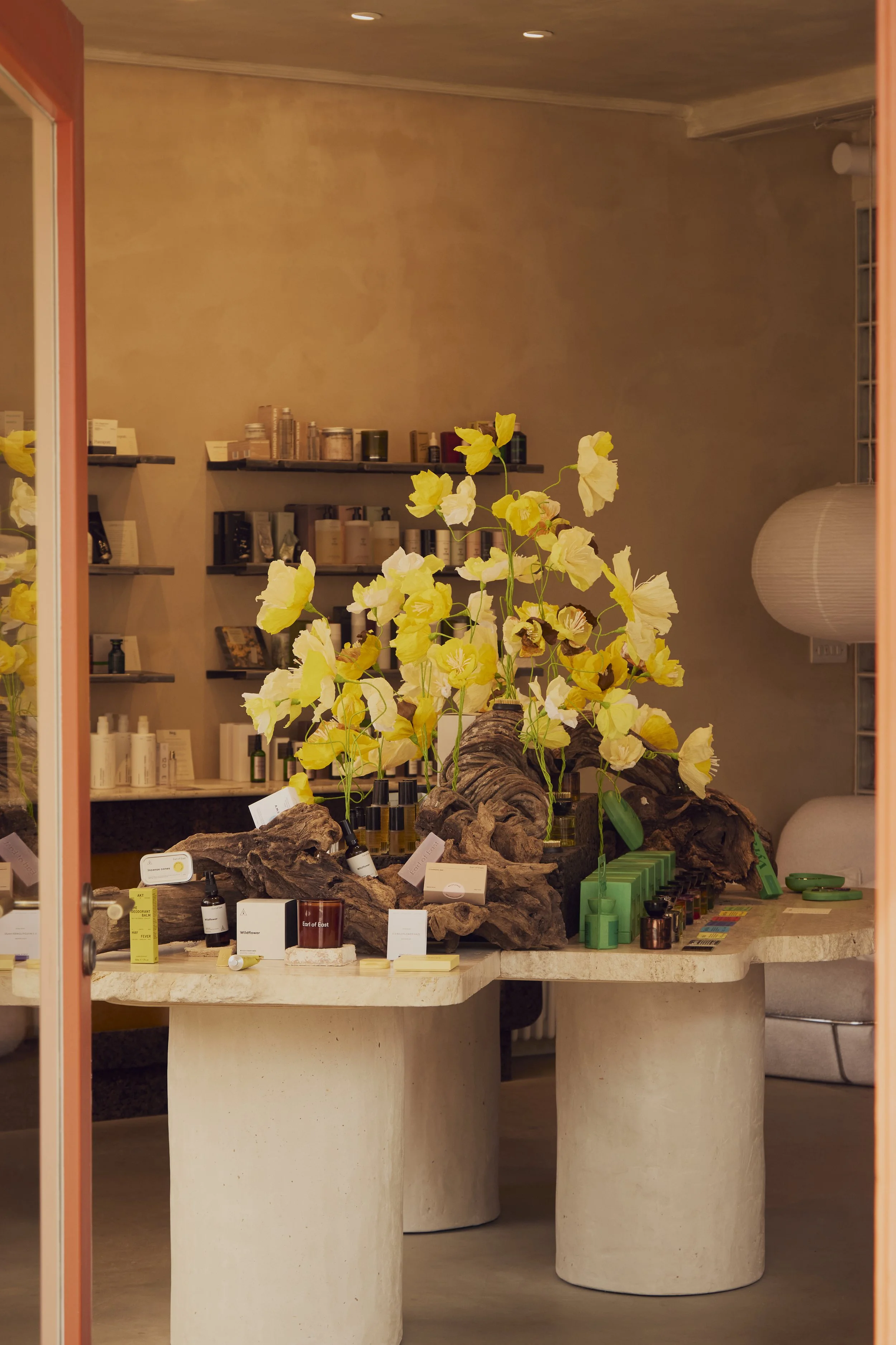 A display table with yellow and white artificial flowers, skincare products, and small labeled signs in a boutique or spa setting, reflected in a glass door.