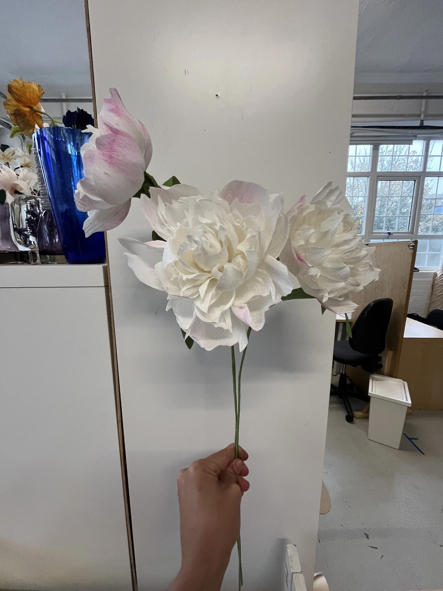Emmeli Kimhi's hand holds a bouquet of large white and pink artificial flowers in front of a plain white wall, with a room and windows visible in the background.
