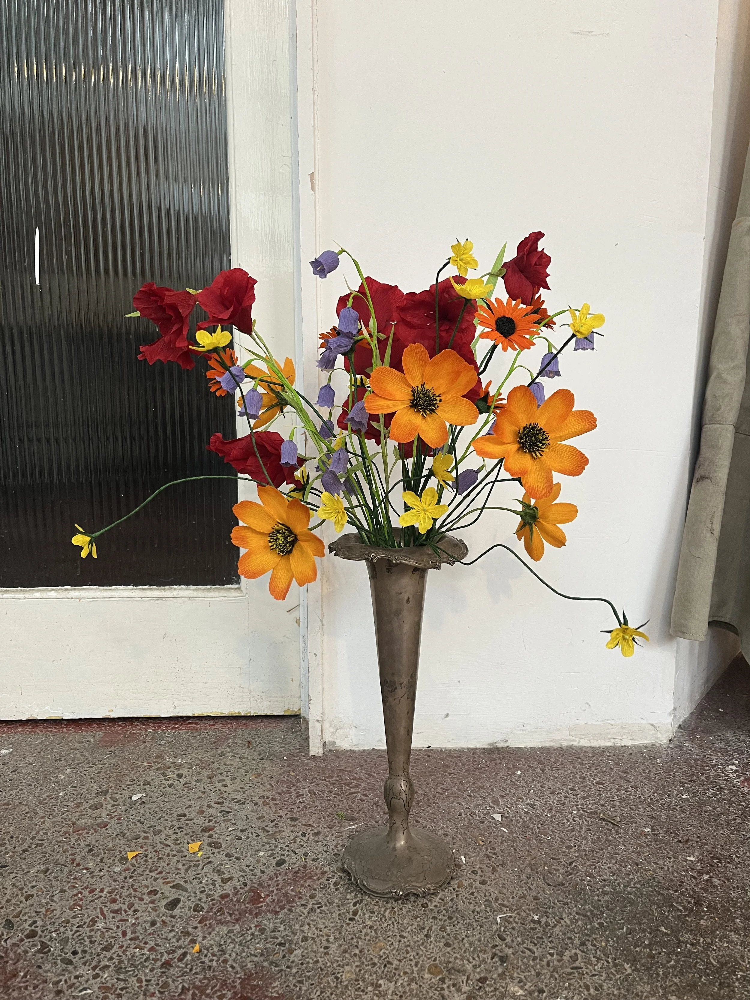 A tall, ornate metal vase with a narrow base and wide opening, filled with a bouquet of colorful paper flowers by Emmeli Kimhi including orange, yellow, red, and purple blooms, placed on a textured concrete floor in front of a white wall 