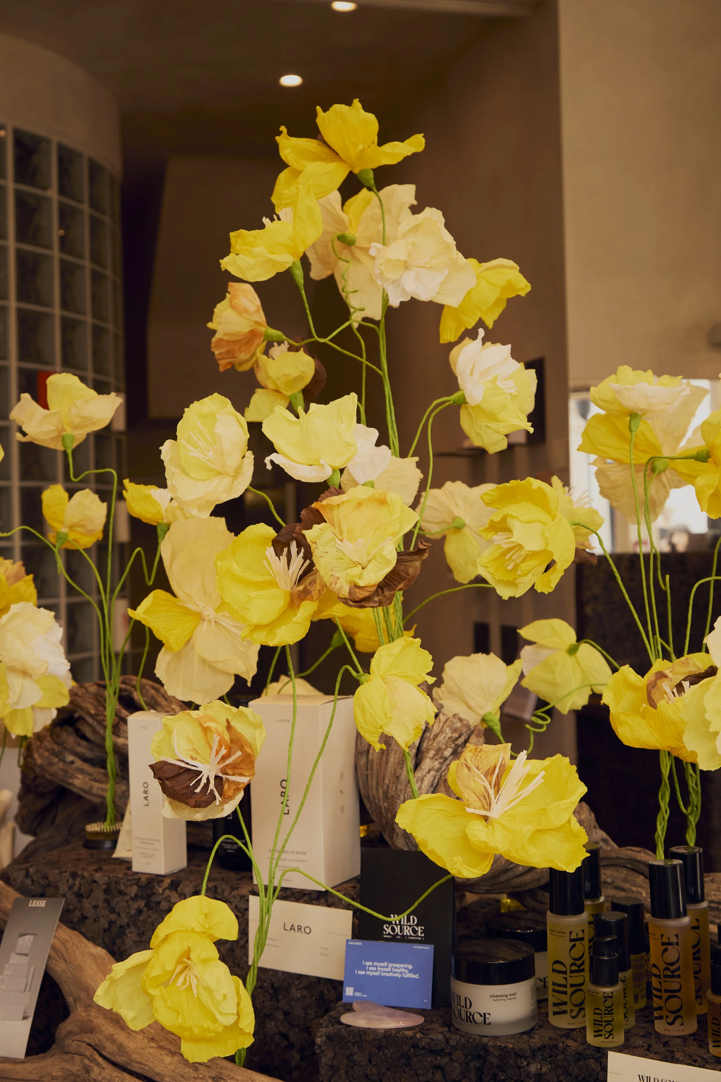 Display of yellow artificial flowers arranged among product bottles and boxes on a wooden surface inside a store.