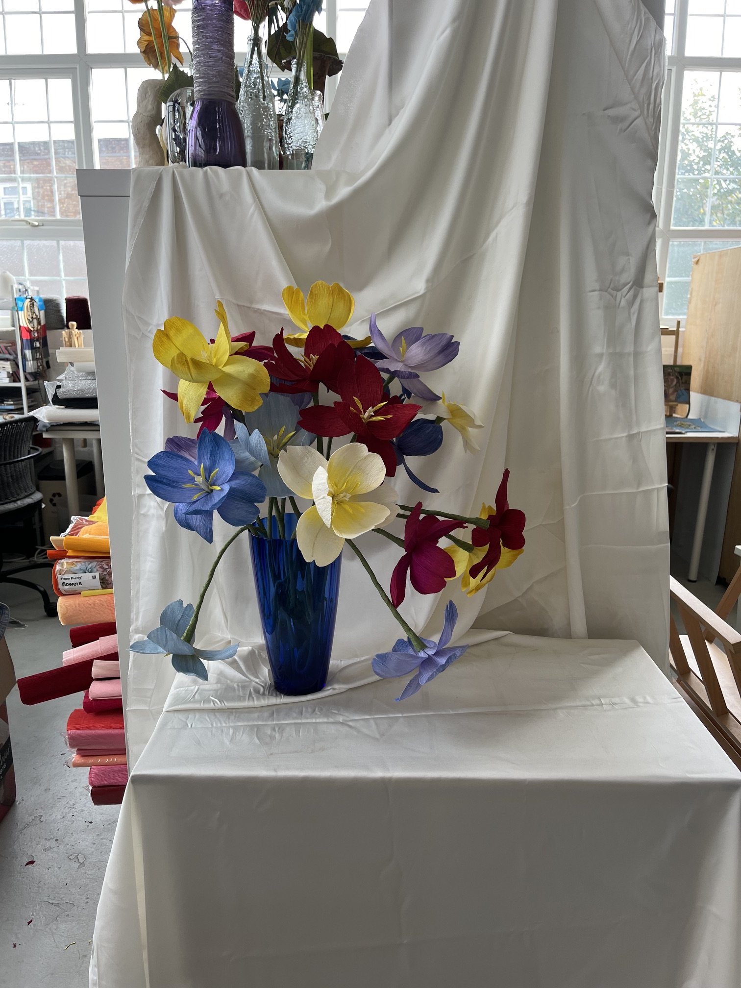A floral arrangement by Emmeli Kimhi in a tall dark blue glass vase with yellow, red, purple, and blue artificial flowers and leaves. The background shows a room with large windows, white counters, shelves with various items,.