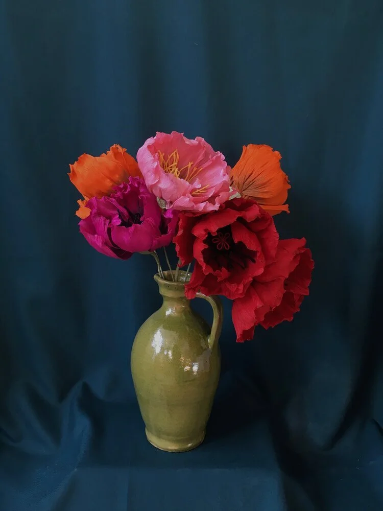 A green ceramic vase with a small handle, holding a bouquet of colorful paper flowers by Emmeli Kimhi including pink, red, orange, and purple, against a dark blue fabric background.
