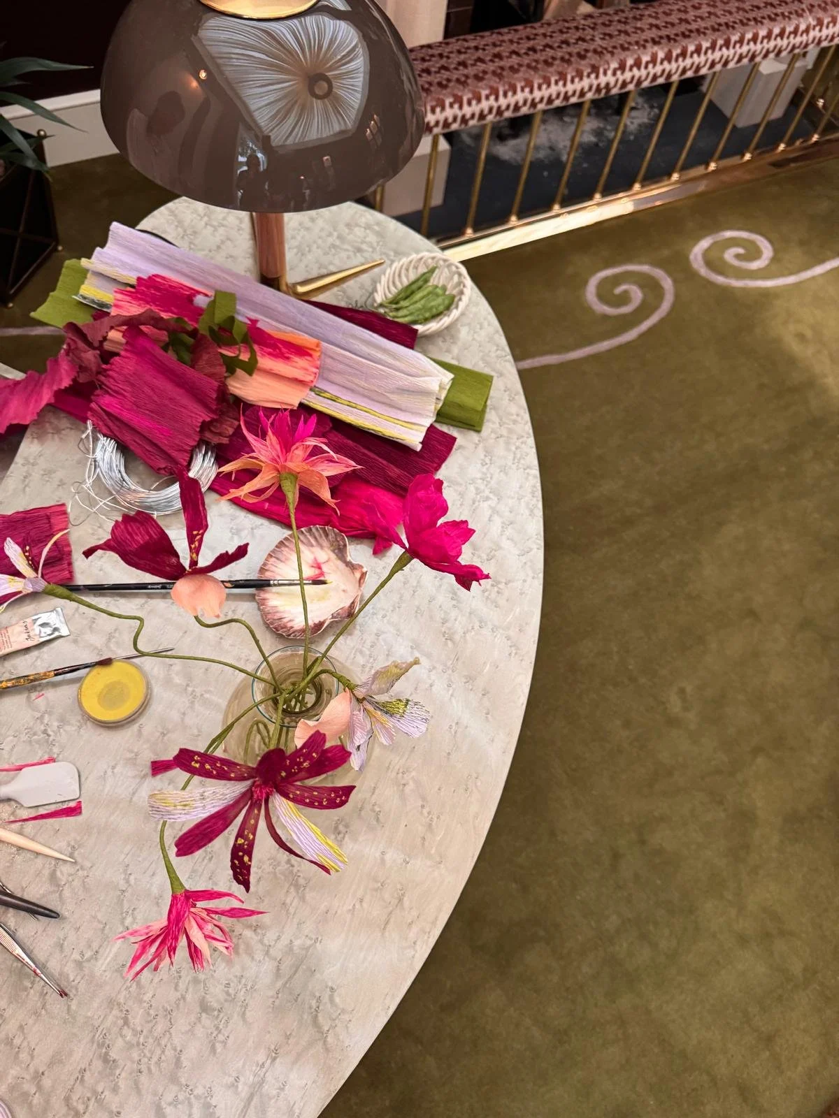 Handcrafted paper flowers displayed in a glass vase alongside hand-dyed crepe papers, painting tools and floral-making materials on a sculptural stone table at the Studio Duggan Speakeasy Salon during WOW!house London.
