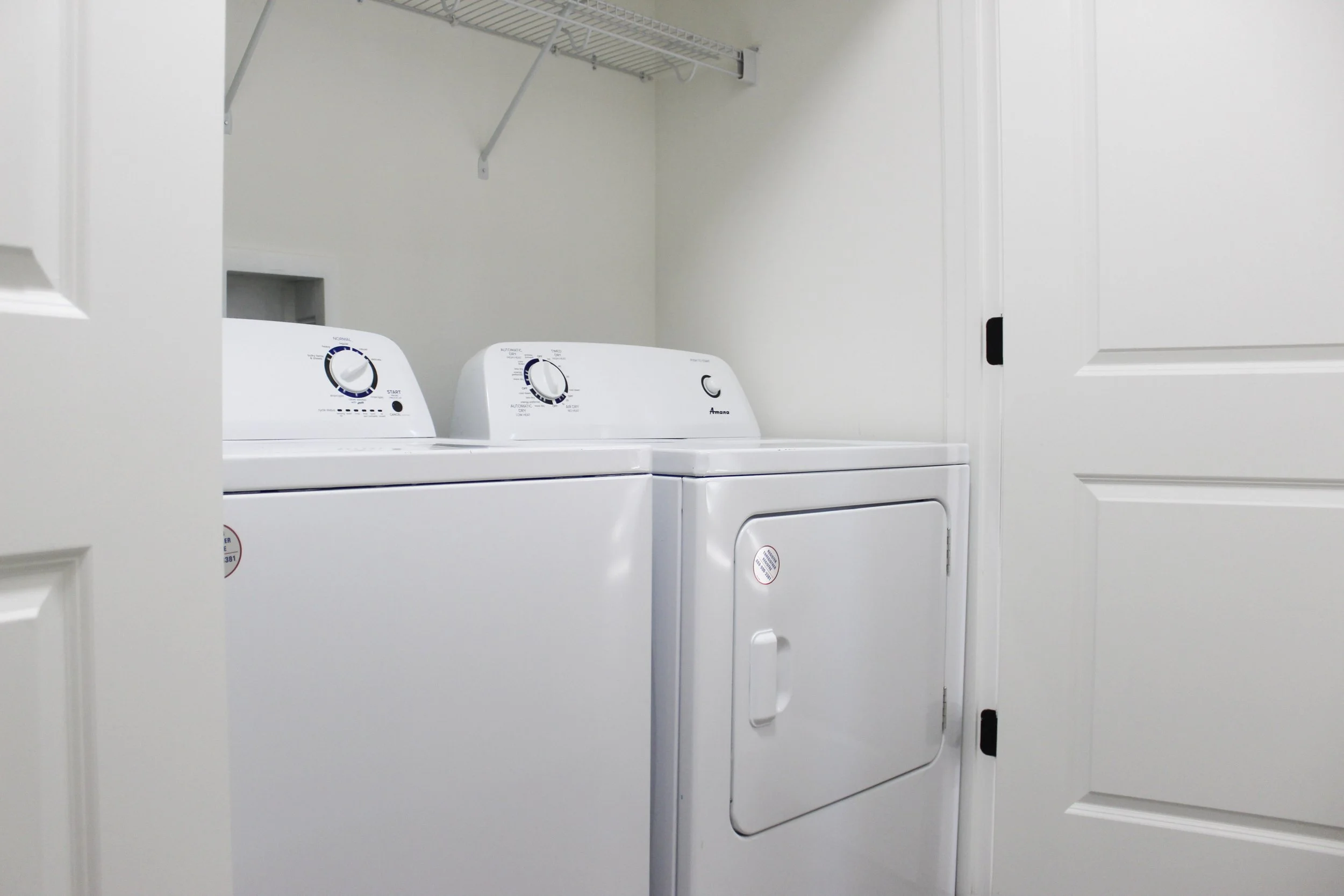 A small laundry room with white washing machine and dryer, with a wire shelf above them, seen through a partially open white door.