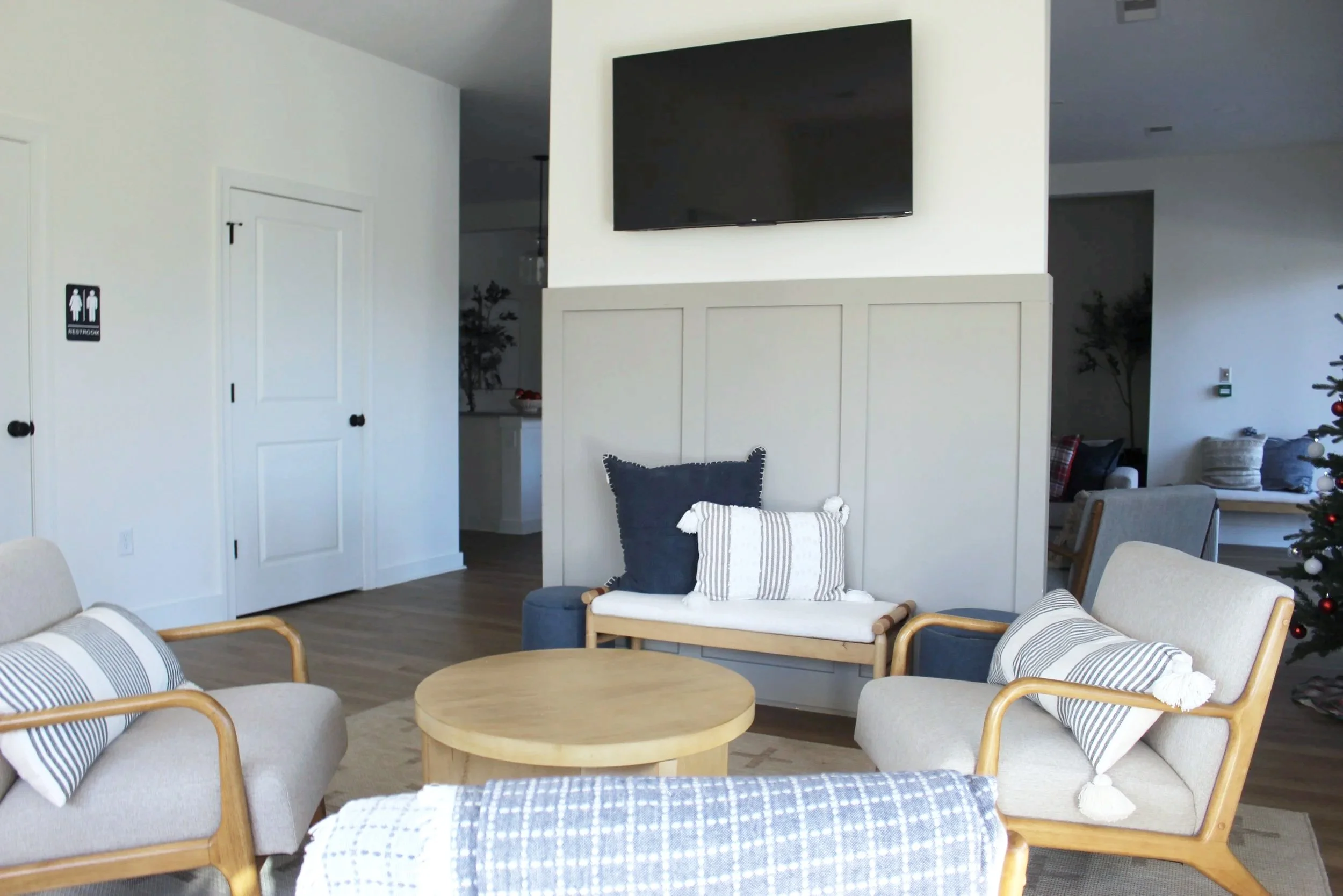 Living room with beige and blue furniture, a round wooden coffee table, pillows with stripes, and a TV mounted on the wall.