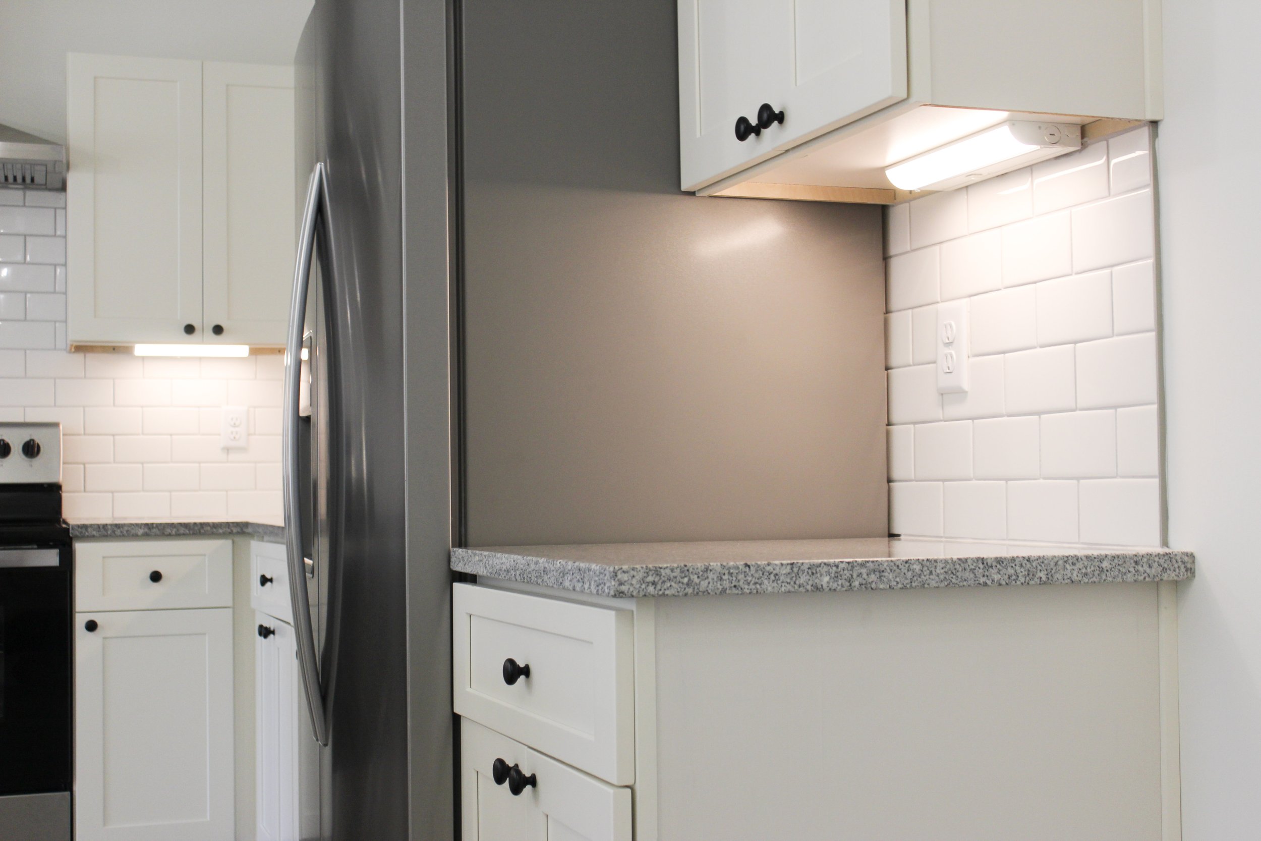 Modern kitchen with white cabinets, granite countertops, and a stainless steel refrigerator. There is a white subway tile backsplash and under-cabinet lighting.