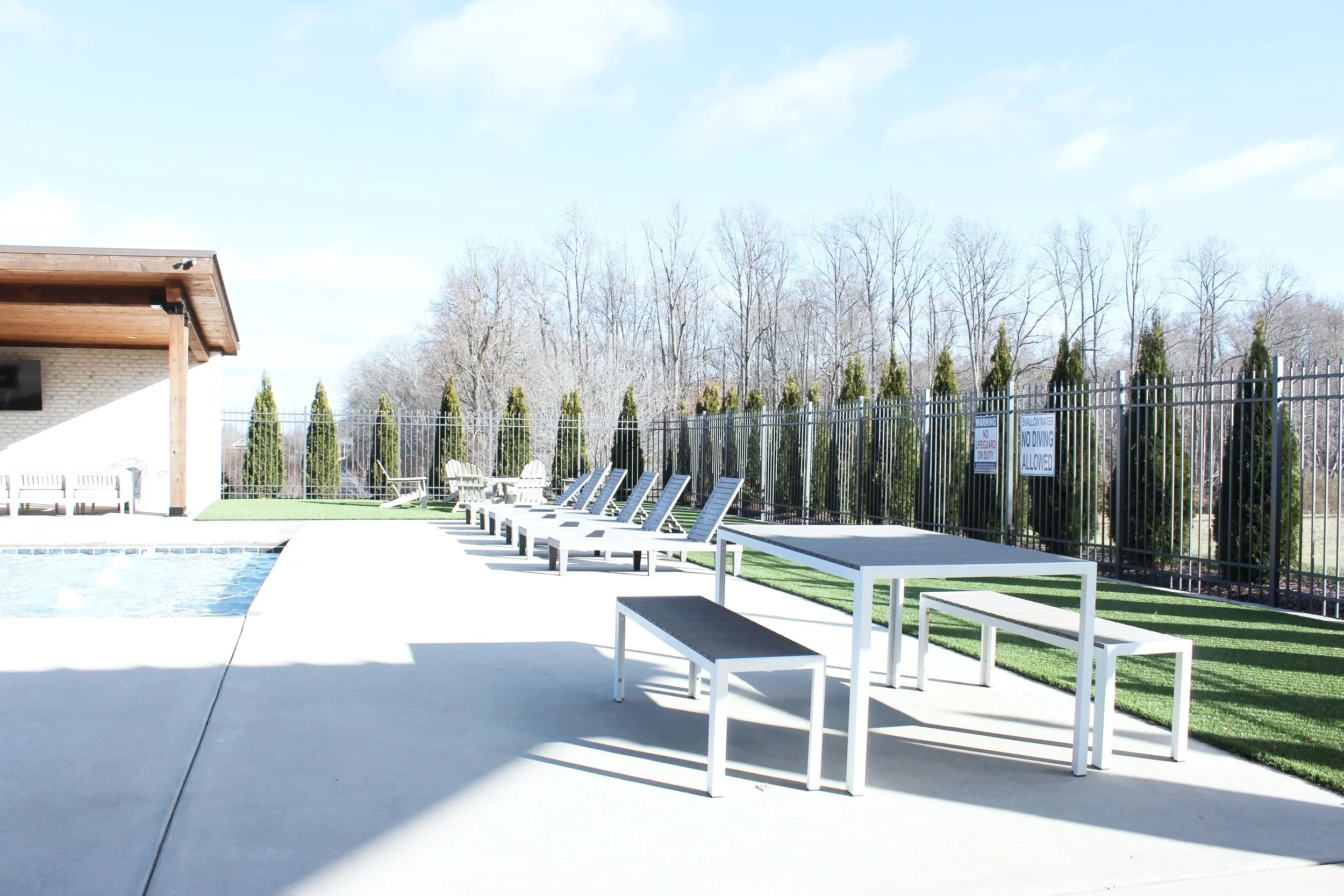 Poolside area with white tables and lounge chairs on a sunny day, surrounded by a black metal fence and trees in the background.