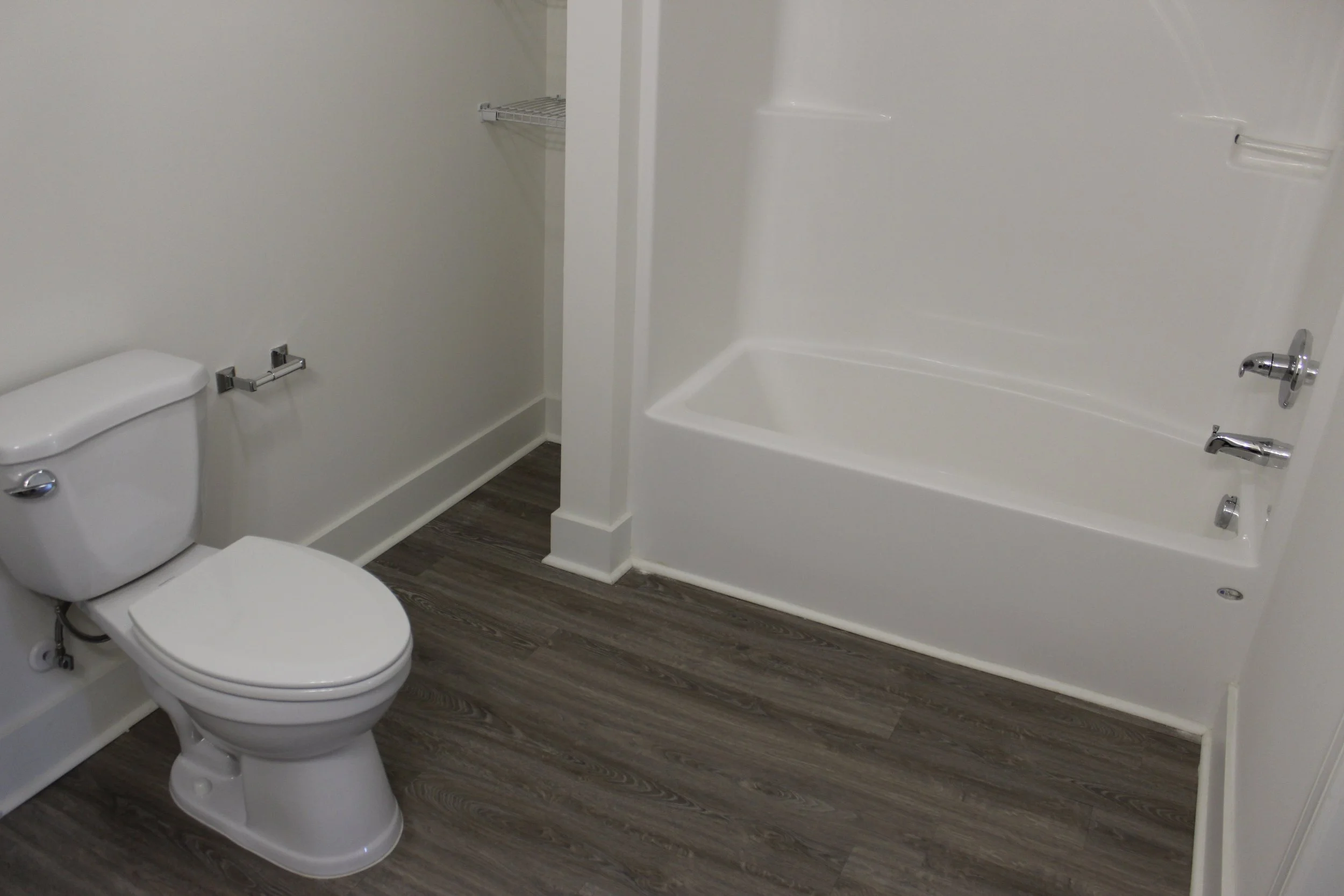 Bathroom with white toilet, empty toilet paper holder, and white bathtub with silver fixtures, dark wood flooring, and white walls.