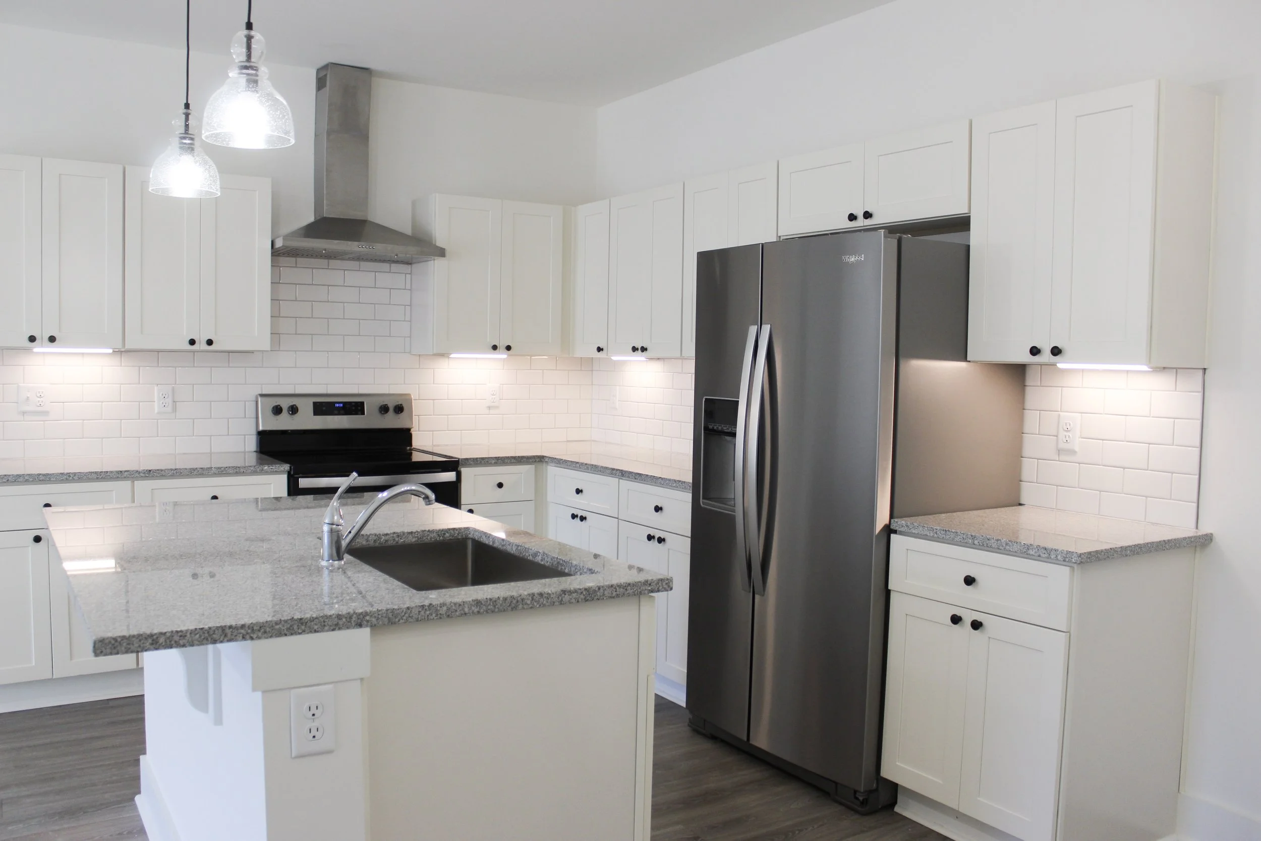 Modern kitchen with white cabinets, granite countertops, stainless steel refrigerator, black stove, white subway tile backsplash, and gray wood flooring.