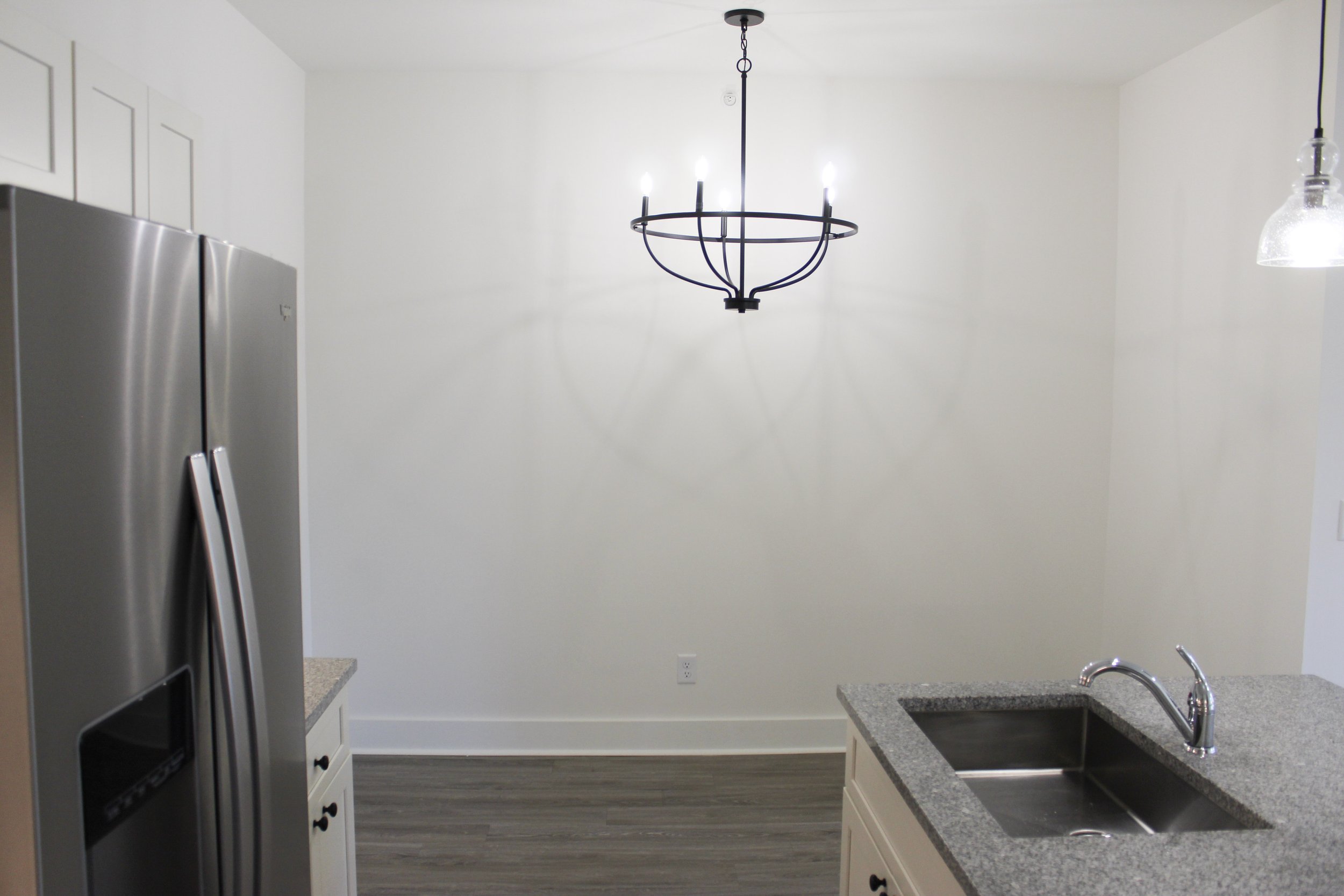 Empty white kitchen with a gray granite countertop island, stainless steel double door refrigerator on the left, stainless steel sink with a chrome faucet on the right, hardwood flooring, and a modern black chandelier hanging from the ceiling.