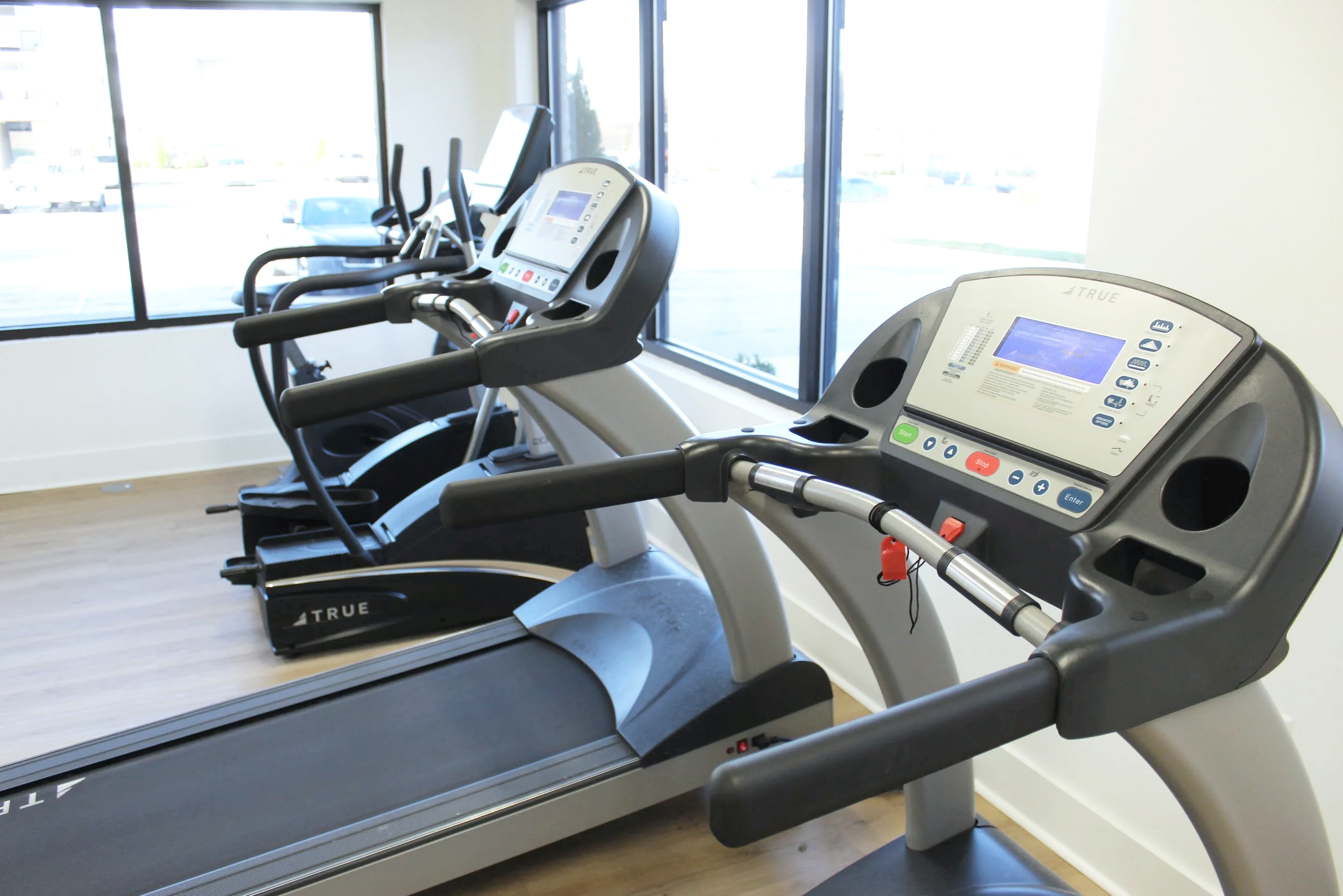 Two treadmills in a gym room near large windows, with a view of parked cars outside.