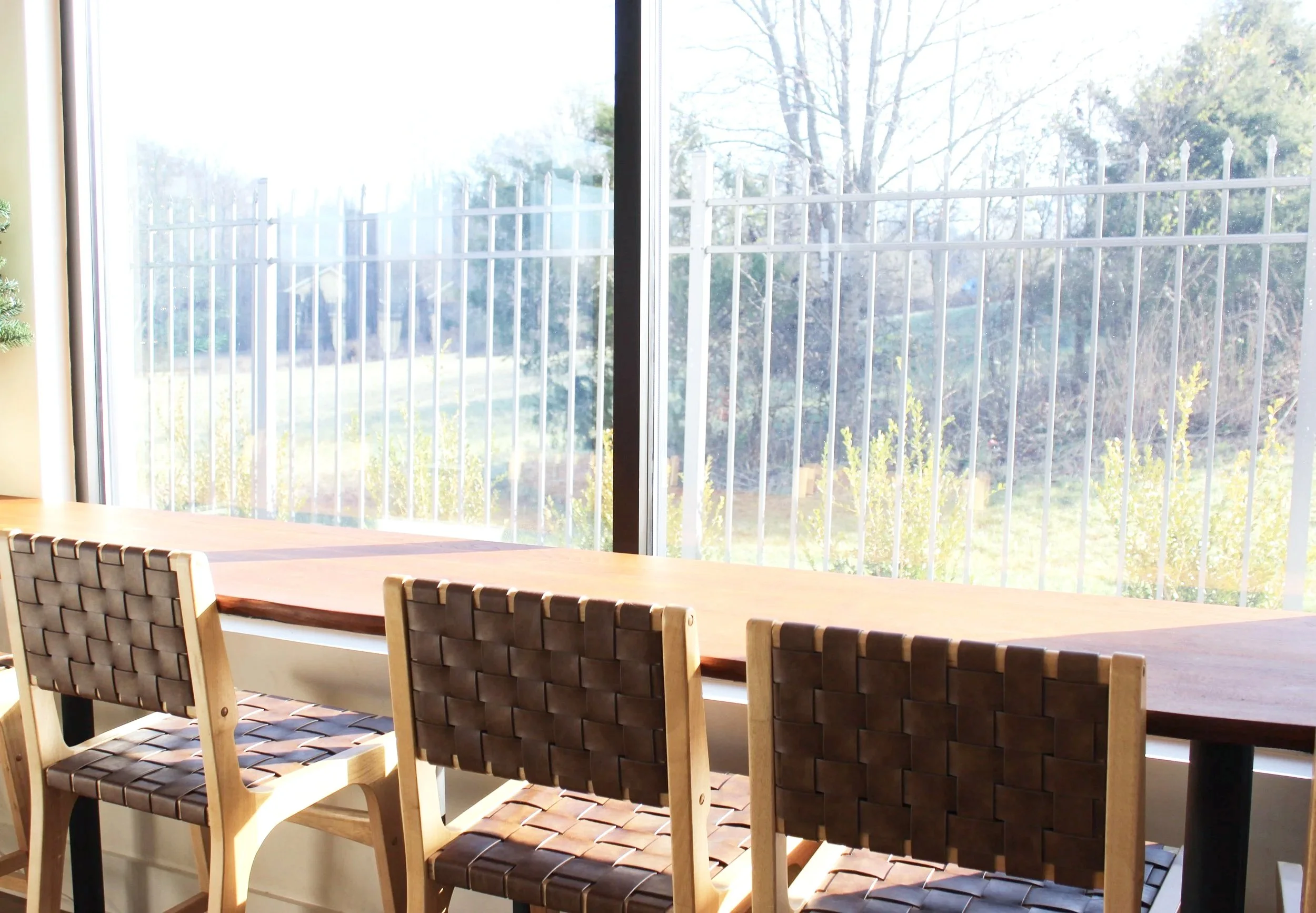 Wooden dining table with four woven leather chairs, large glass window revealing a fenced outdoor area with trees and bushes.