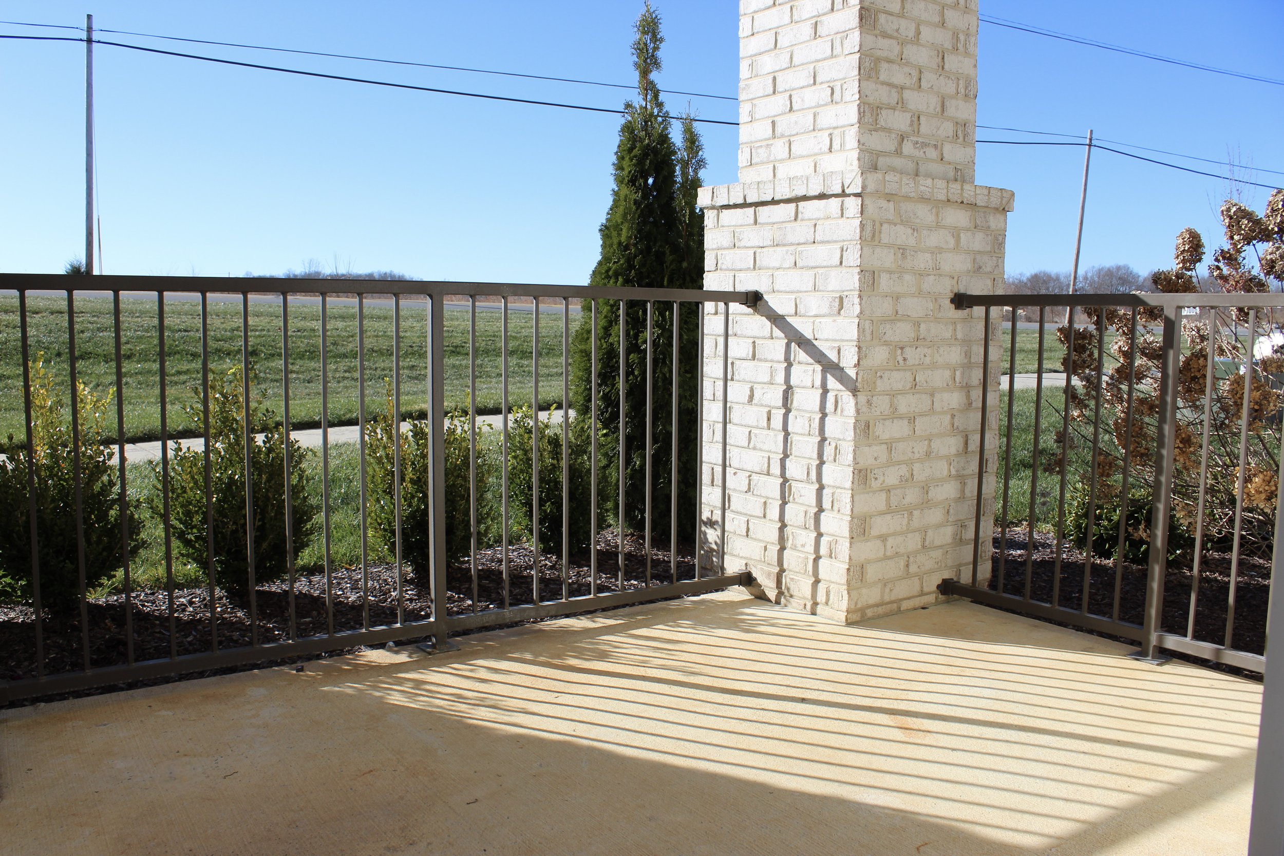 A porch with black metal railings, a brick column, and a view of a grassy area with a sidewalk and bushes, under a clear blue sky.