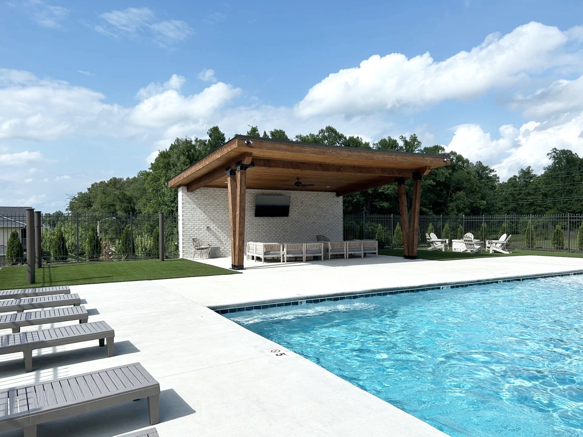 A swimming pool with lounge chairs along the side and a shaded cabana area with outdoor chairs and a TV, surrounded by a black fence, under a partly cloudy sky.
