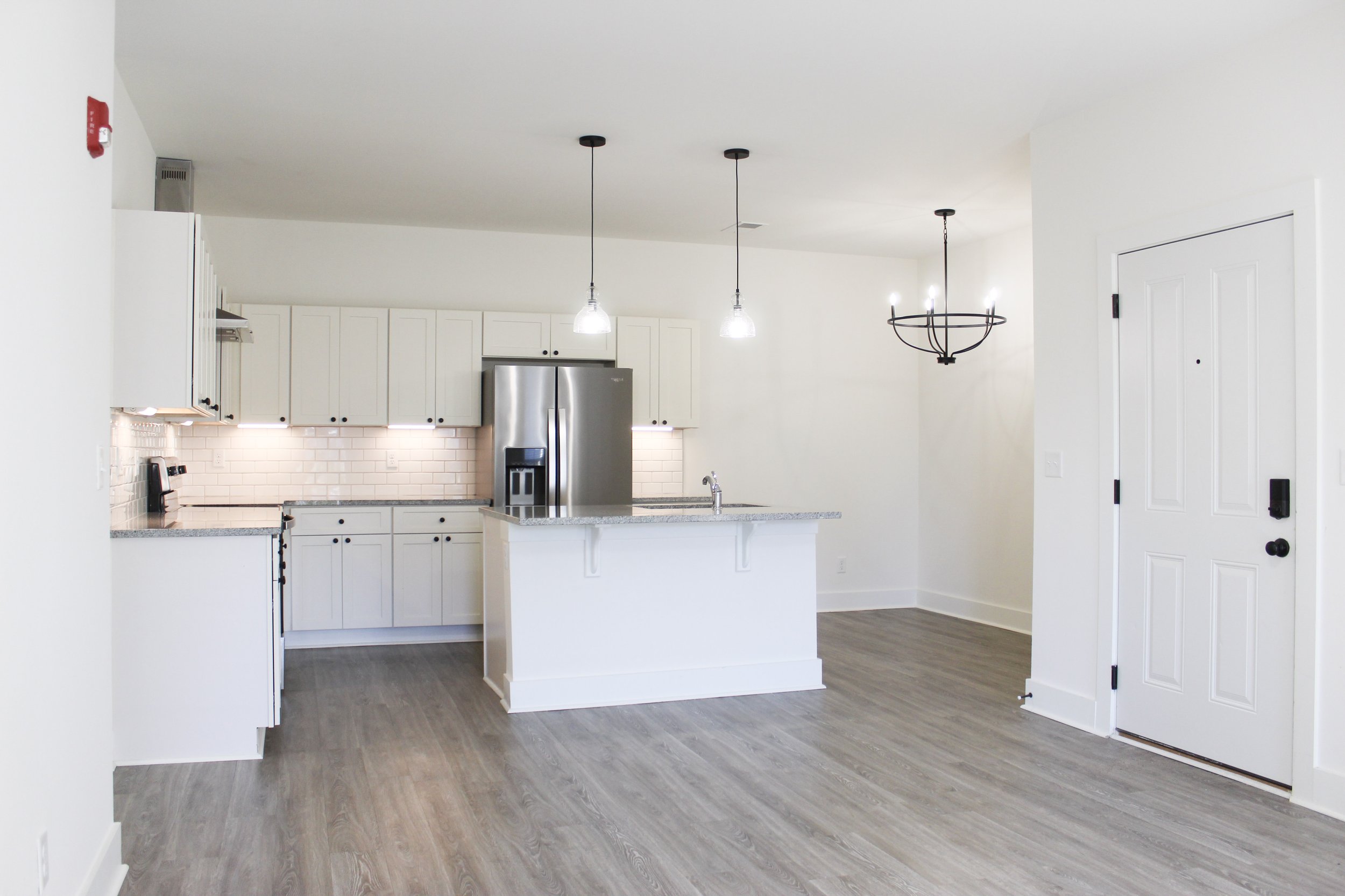 Empty modern kitchen with white cabinets, stainless steel refrigerator, granite countertops, white backsplash, pendant lights, and a round black chandelier, with a gray wood floor and white walls.