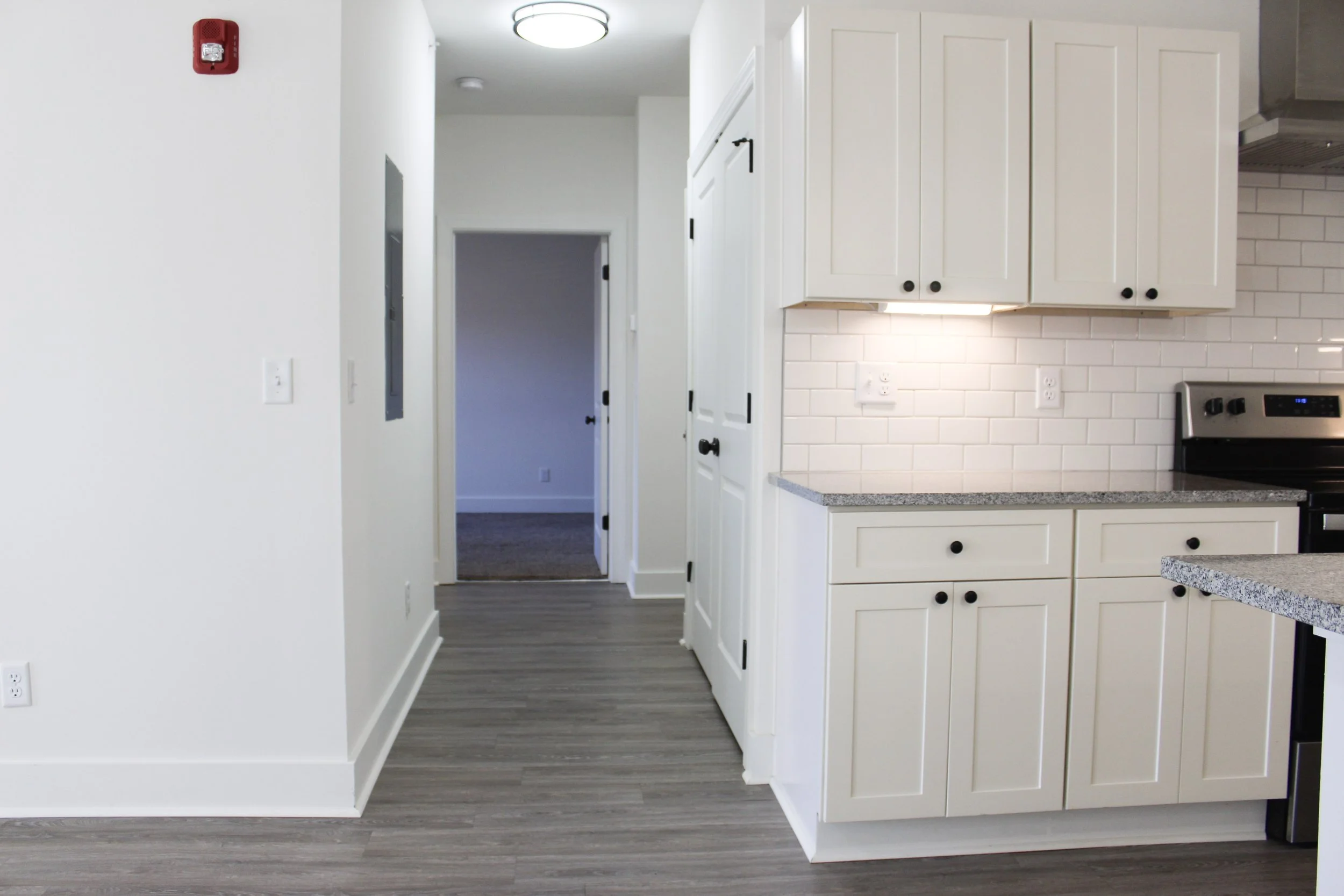 Kitchen with white cabinets, gray countertops, and white subway tile backsplash. Partial view of stove on the right, white walls, and a hallway leading to a room with carpeted floor.