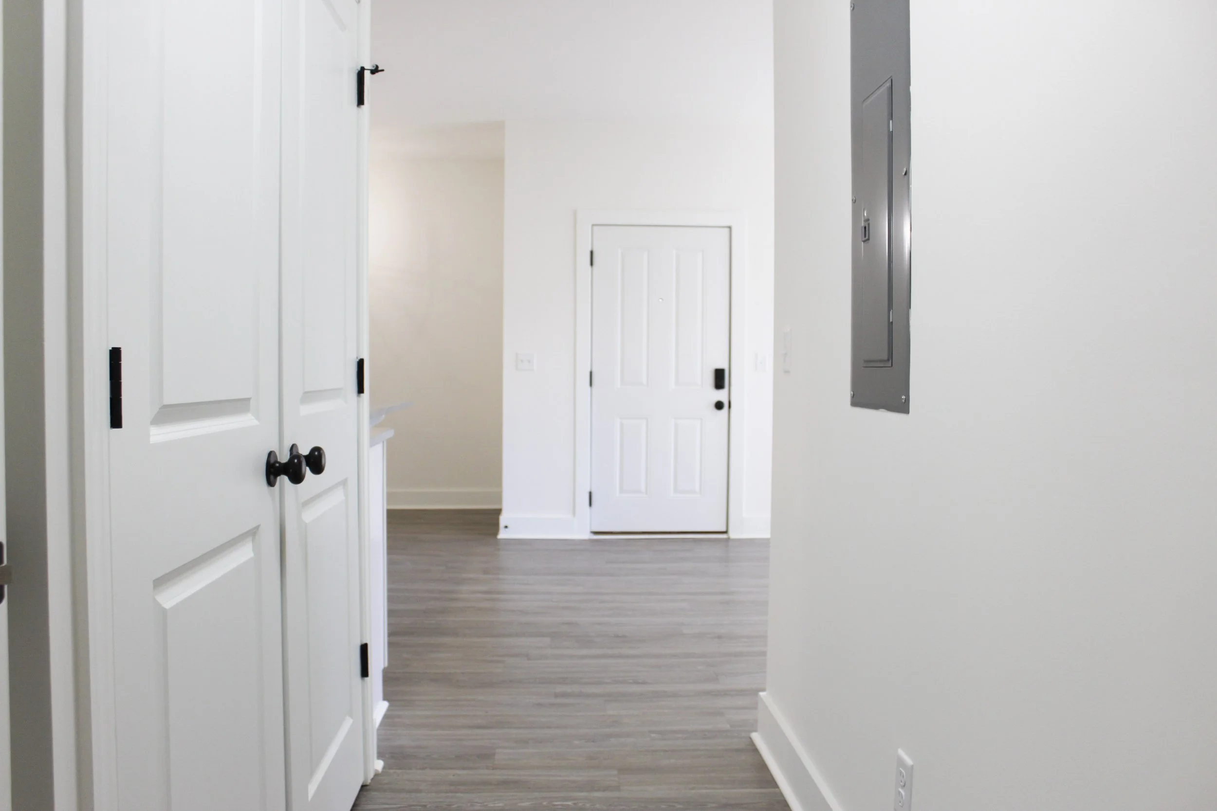 Interior view of a modern apartment hallway with white walls, a closed white front door, and a grey electrical panel on the right wall.