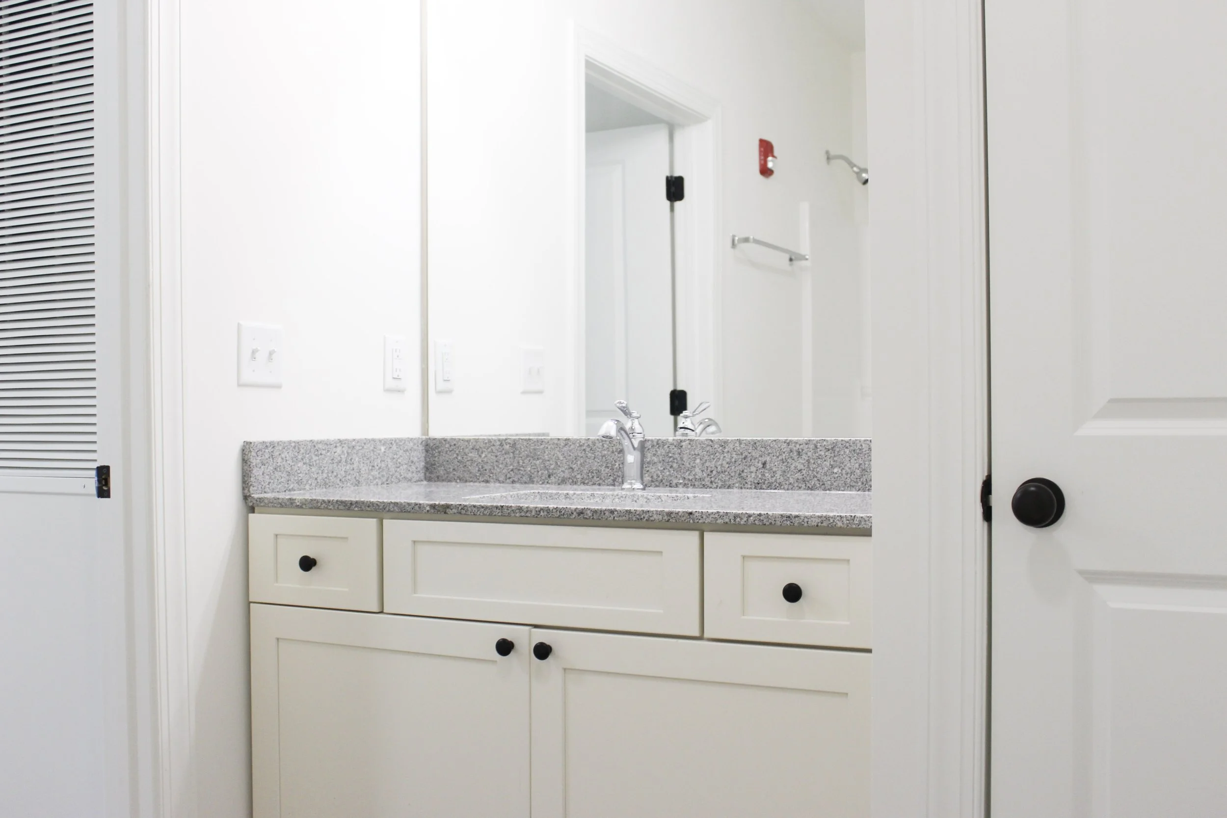Bathroom vanity with a granite countertop and a mirror, with a door slightly open revealing a towel bar and an emergency alarm on the wall.