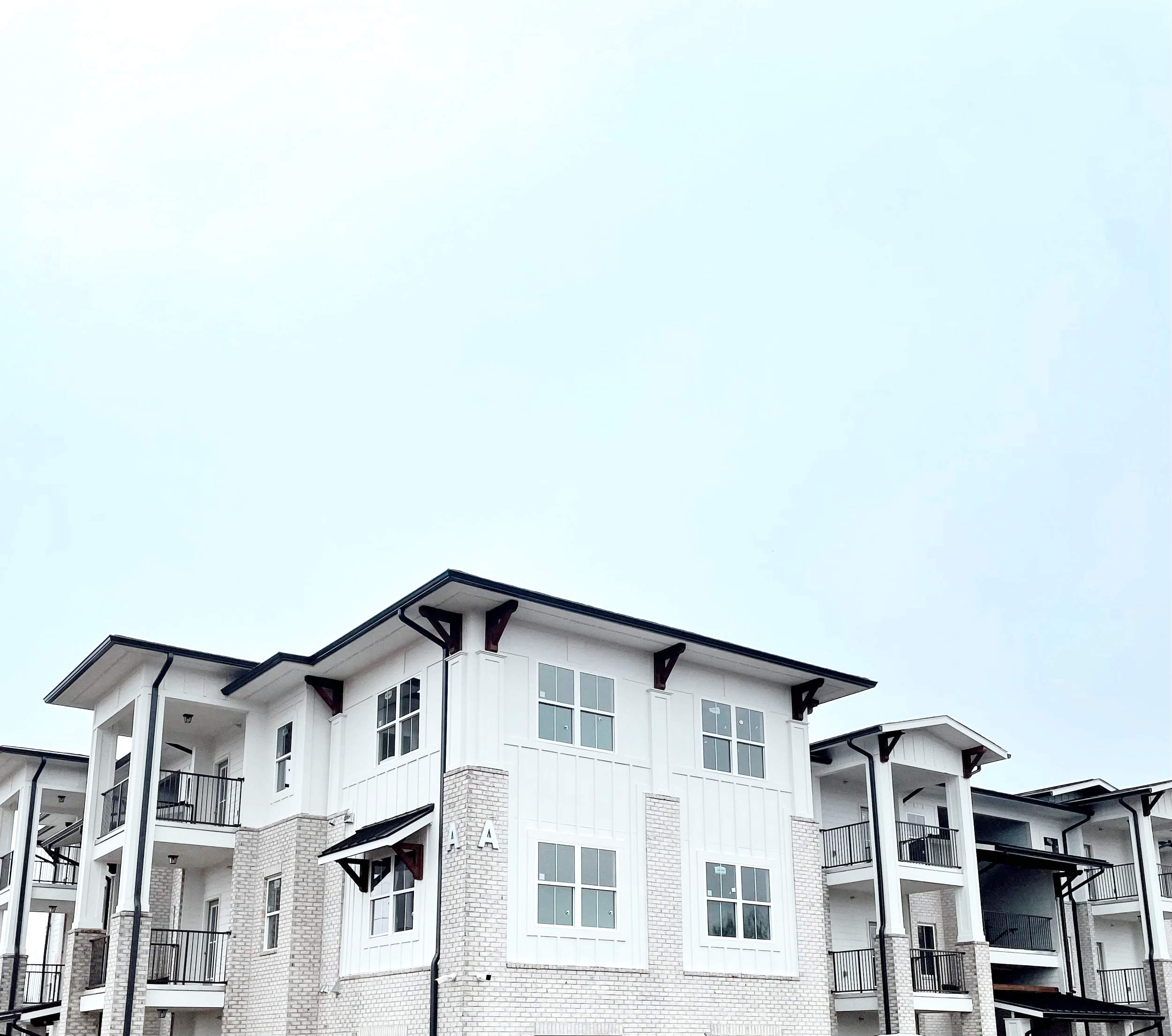 Modern multi-story apartment building with white exterior, black balconies, and large windows under a cloudy sky.