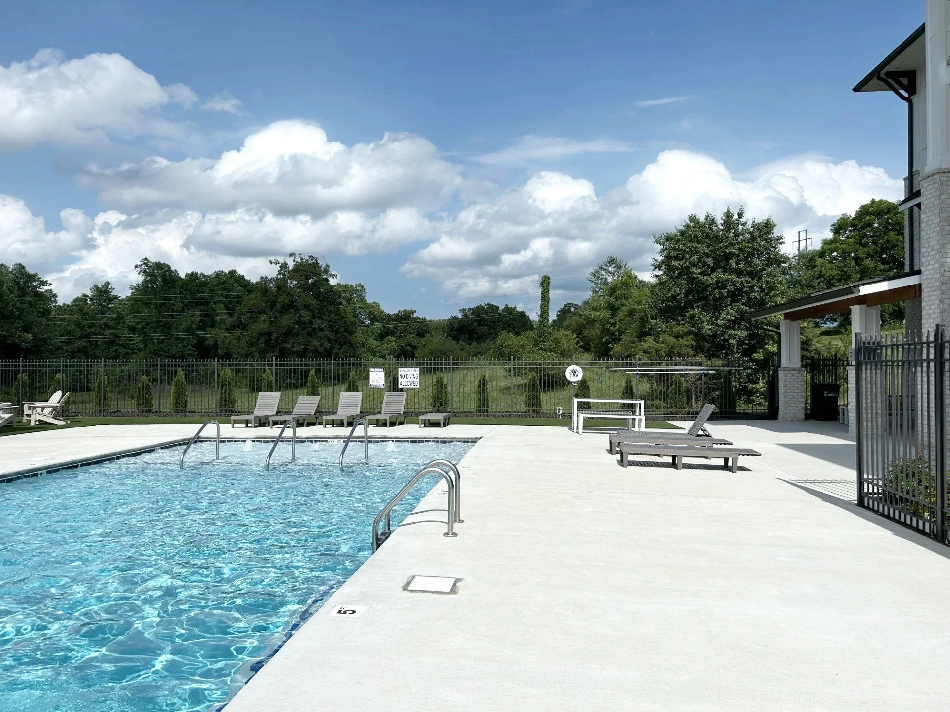 Empty swimming pool on a sunny day with lounge chairs, a building, and trees in the background.