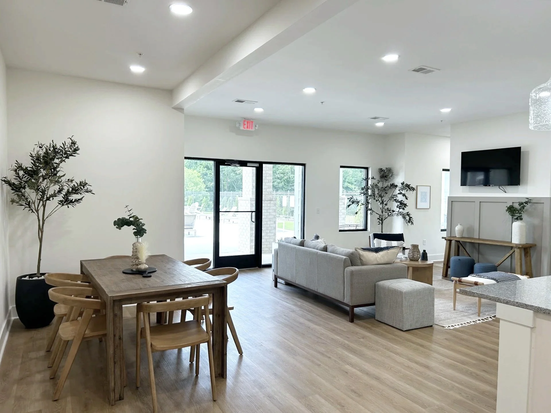 Open-concept living and dining room with wooden flooring, beige walls, large windows, a gray sofa, a wooden dining table with six chairs, potted plants, wall-mounted television, and decorative vases.