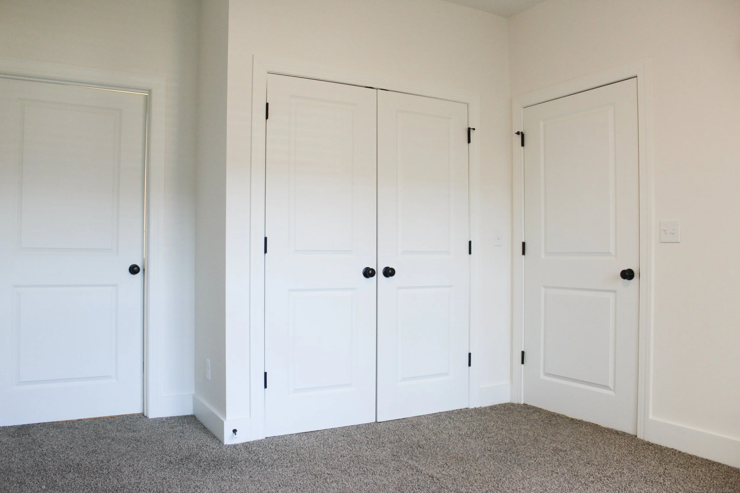 A room with three white closed doors, two with black doorknobs on the right and one with a black doorknob on the left, beige carpeted floor, and white walls.