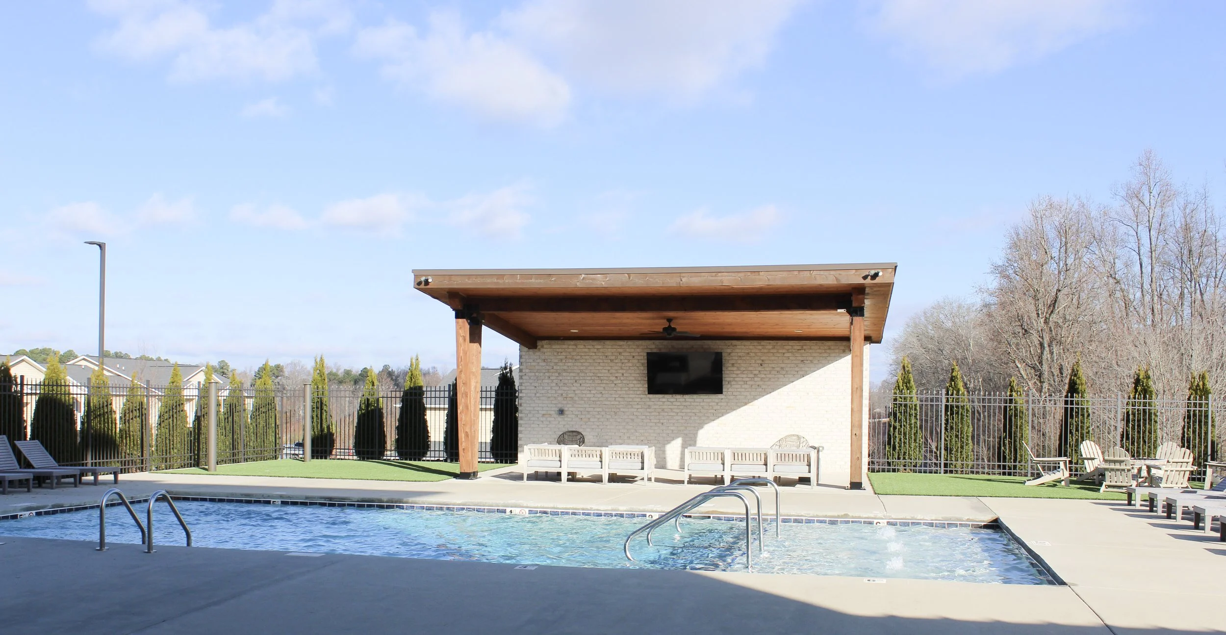 Outdoor pool area with lounge chairs, a covered seating area with a TV on a brick wall, and trees in the background.