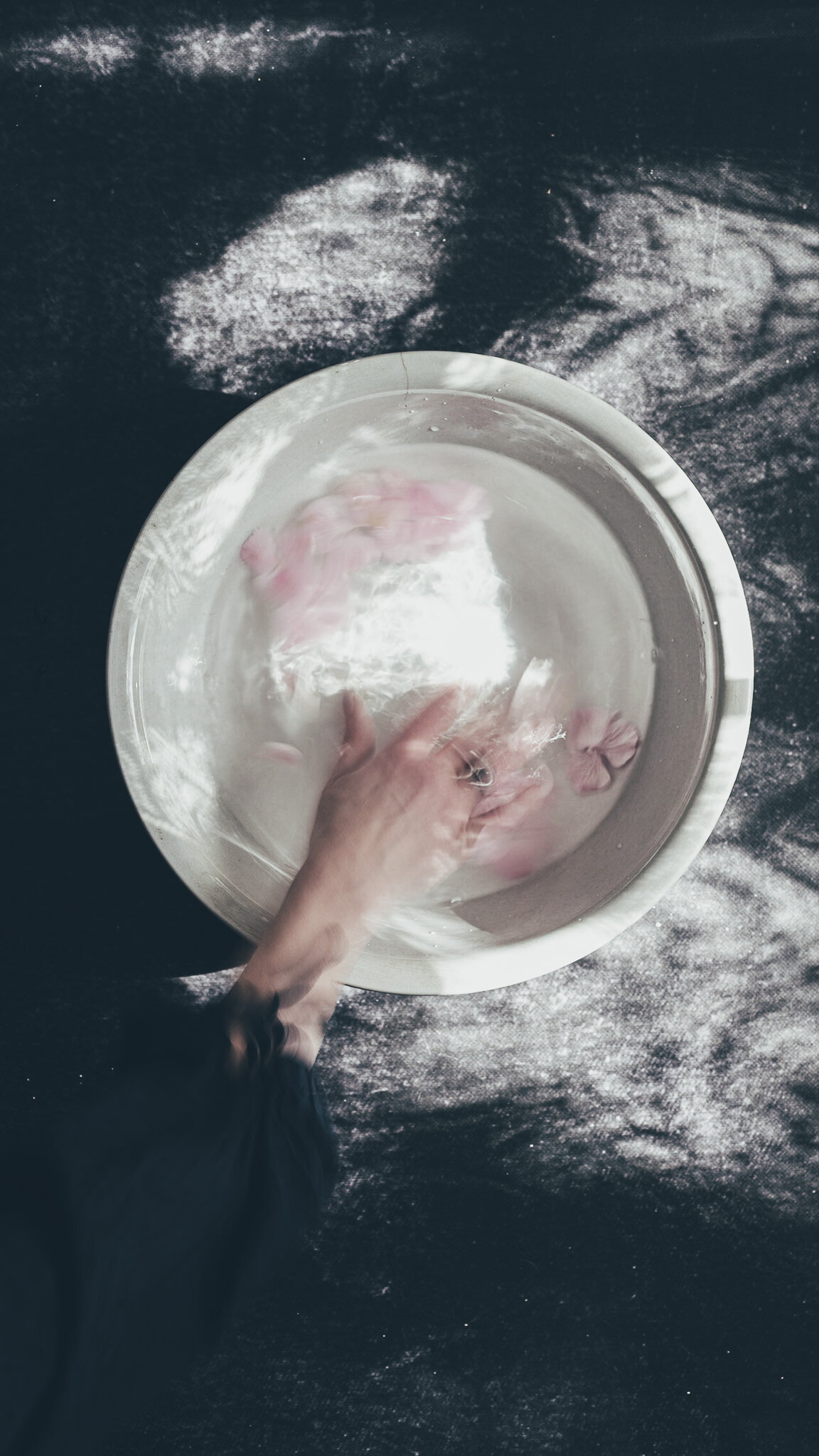 A person mixing pink flower petals in water in a glass bowl on a dark surface with flour scattered around.