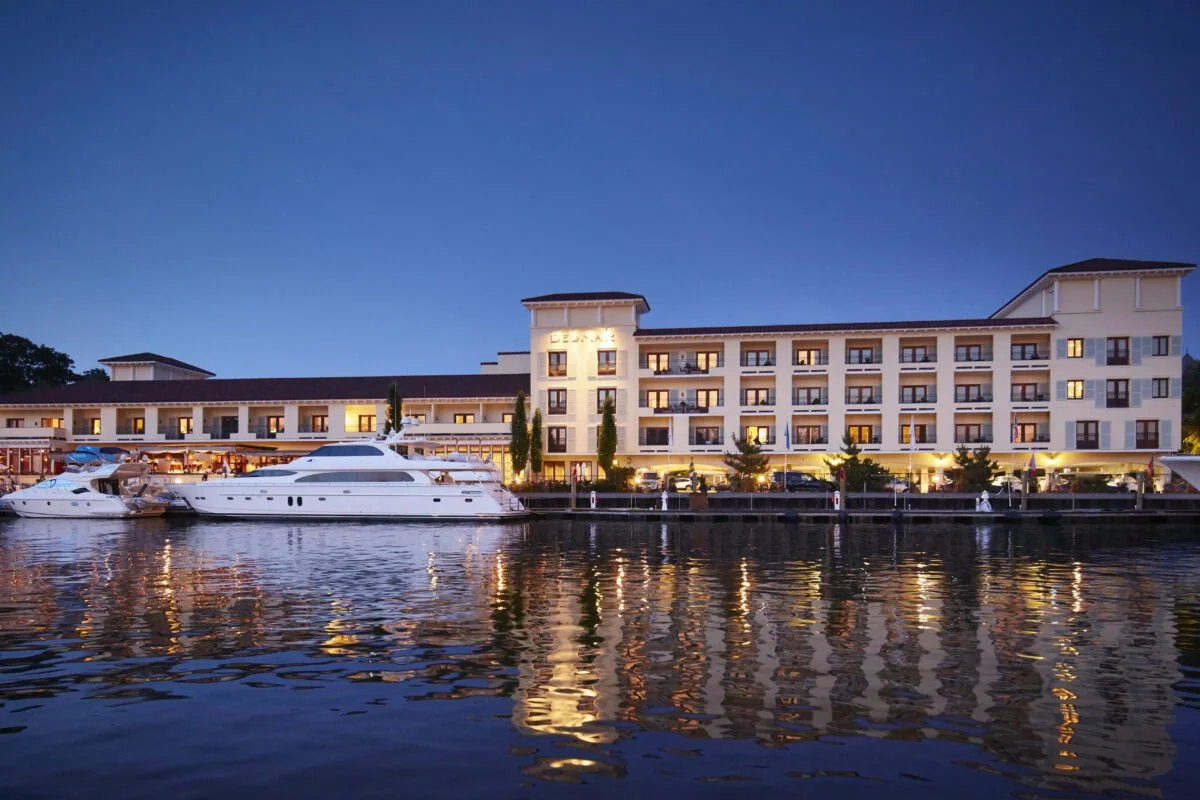 A large hotel or apartment building by a waterfront with boats docked nearby during evening twilight, with the building's lights reflecting on the water.