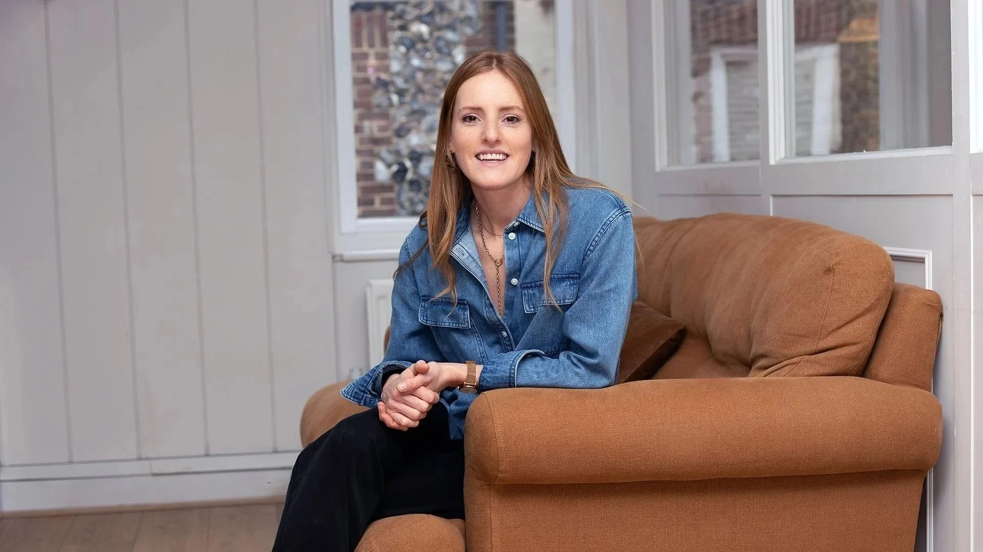 A woman with red hair wearing a denim jacket and black pants sitting on an orange sofa in a room with white panel walls and large windows.