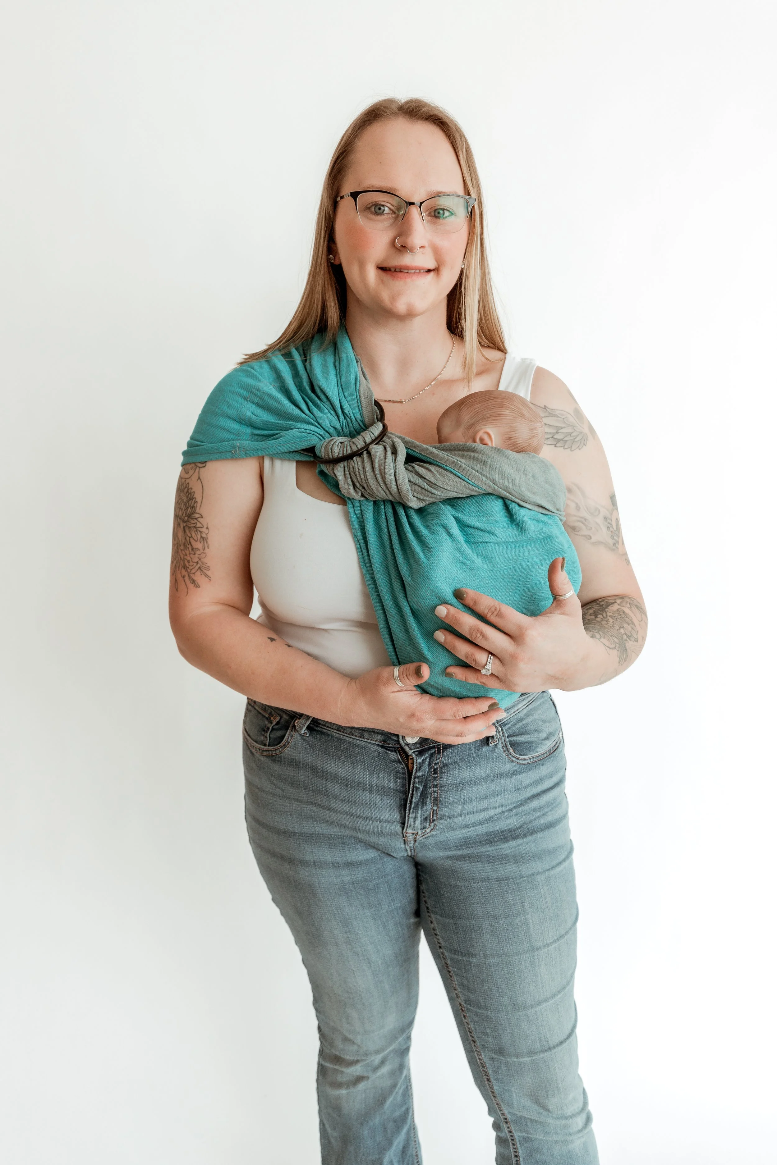 Woman with glasses and tattoos holding a baby in a teal wrap against a white background.