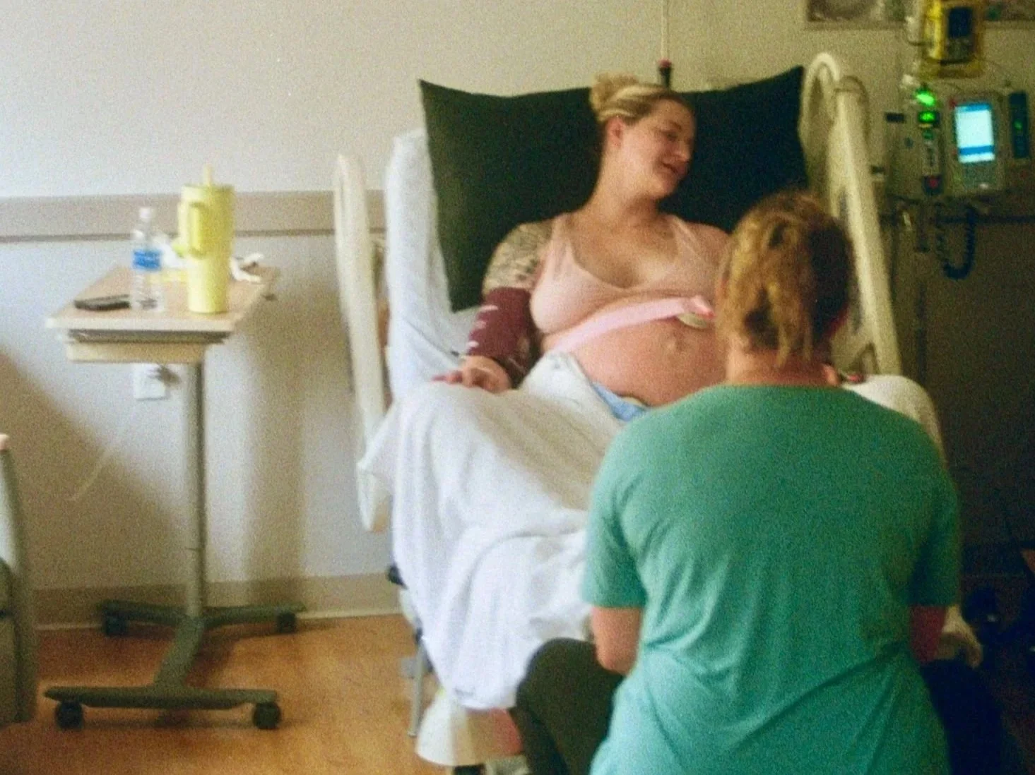 A woman in a hospital bed with a maternity gown, smiling at a person sitting nearby, who is wearing a green shirt, in a hospital room with medical equipment and a bedside table.