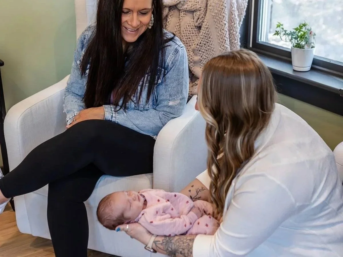 A woman with long hair and a denim jacket sitting on a white armchair, smiling at a young woman with long hair holding a sleeping baby in her lap. The setting is indoors with a large window and a small potted plant on the windowsill.