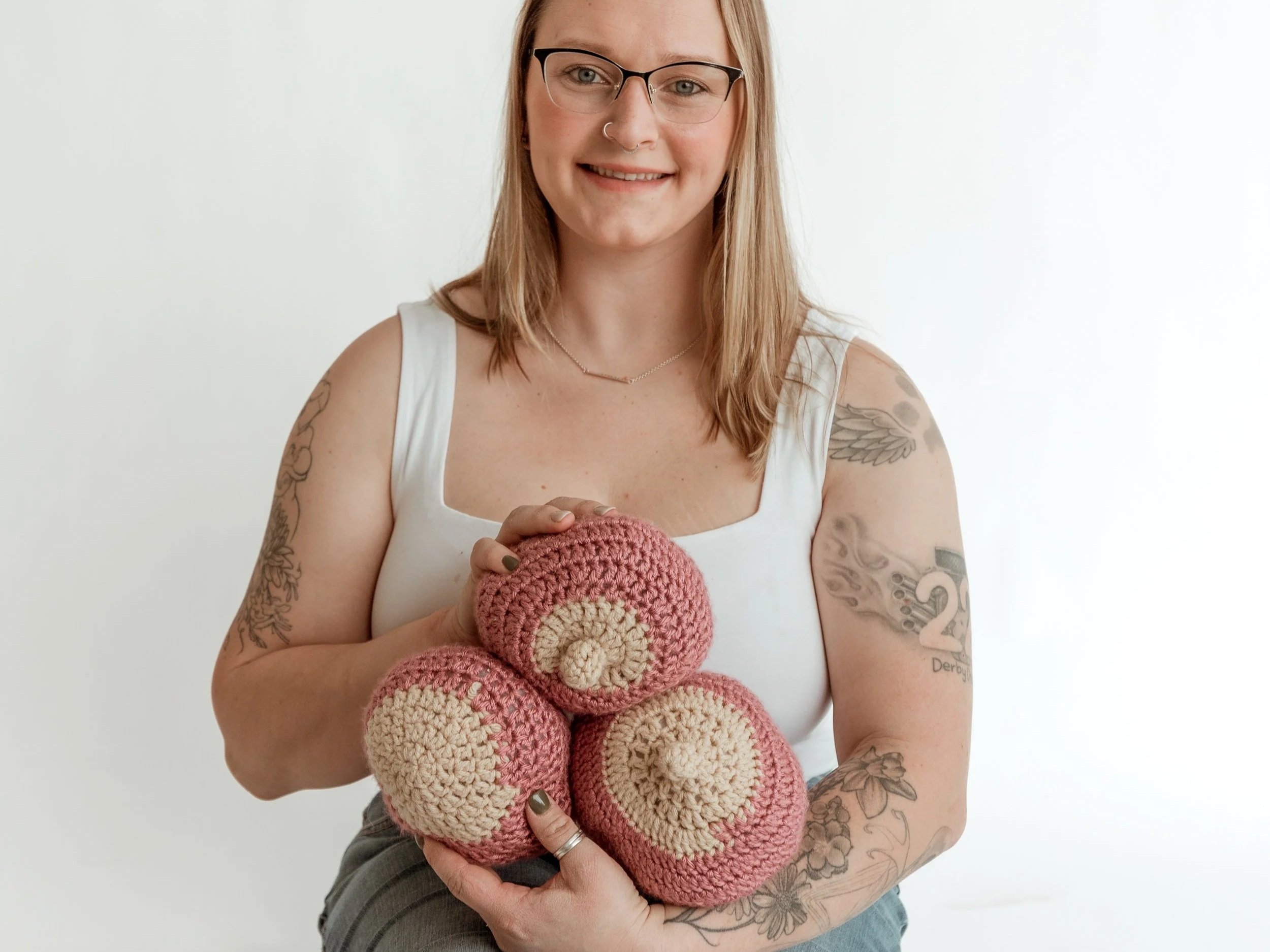 A woman with glasses and tattoos is holding a bunch of pink and beige crocheted baby hats.