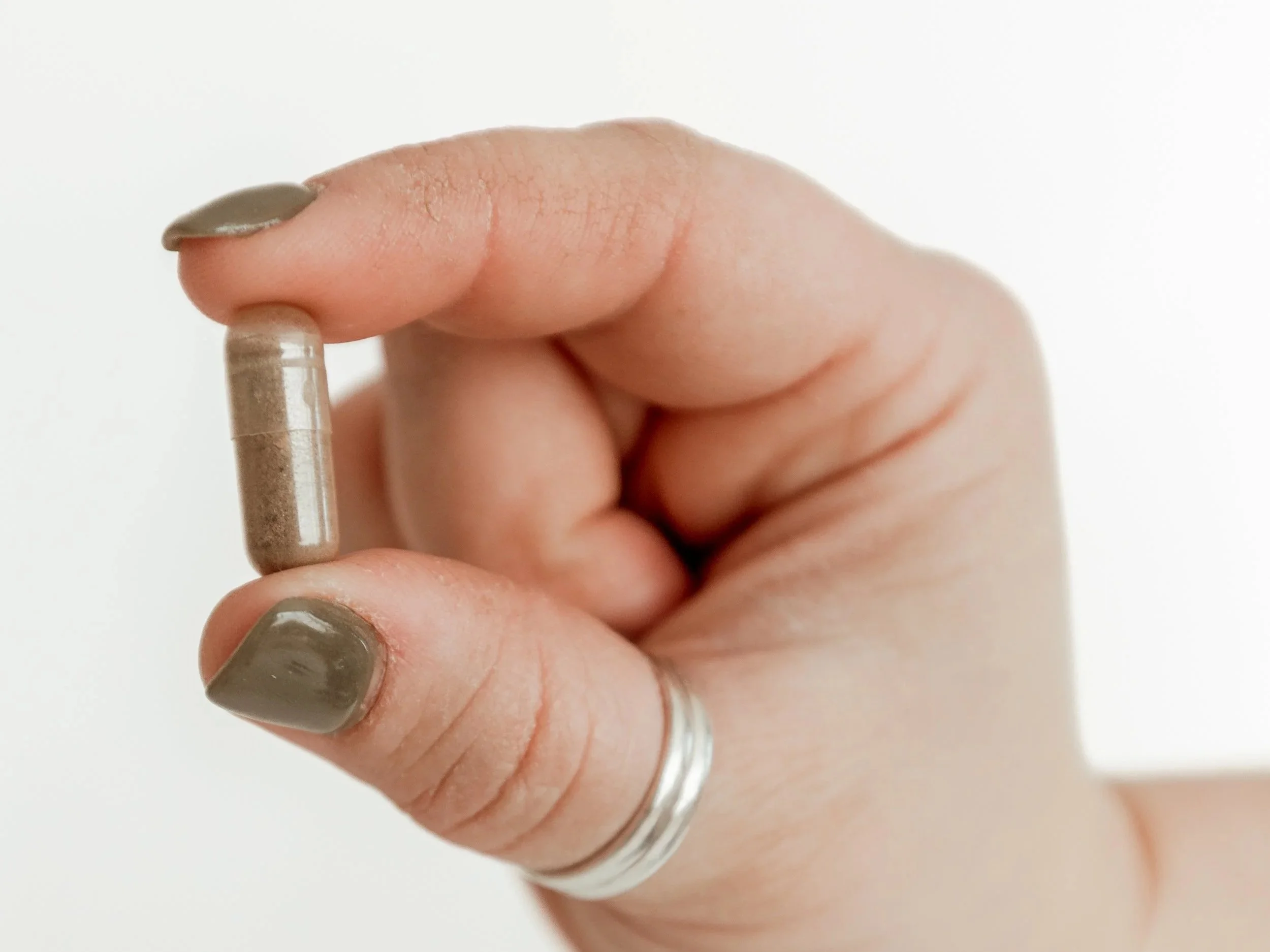 A close-up of a person's hand holding a brown capsule pill, with grayish nail polish on their nails, against a plain white background.