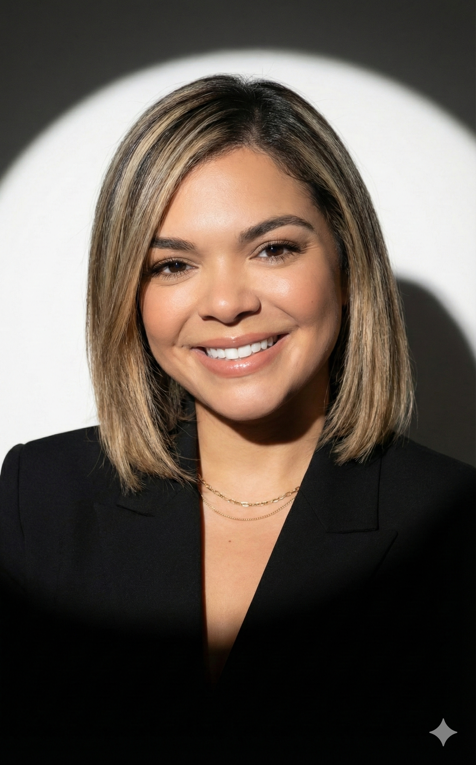 A woman with shoulder-length blonde hair wearing a black blazer and a delicate gold necklace, smiling in front of a white background with a spotlight effect.