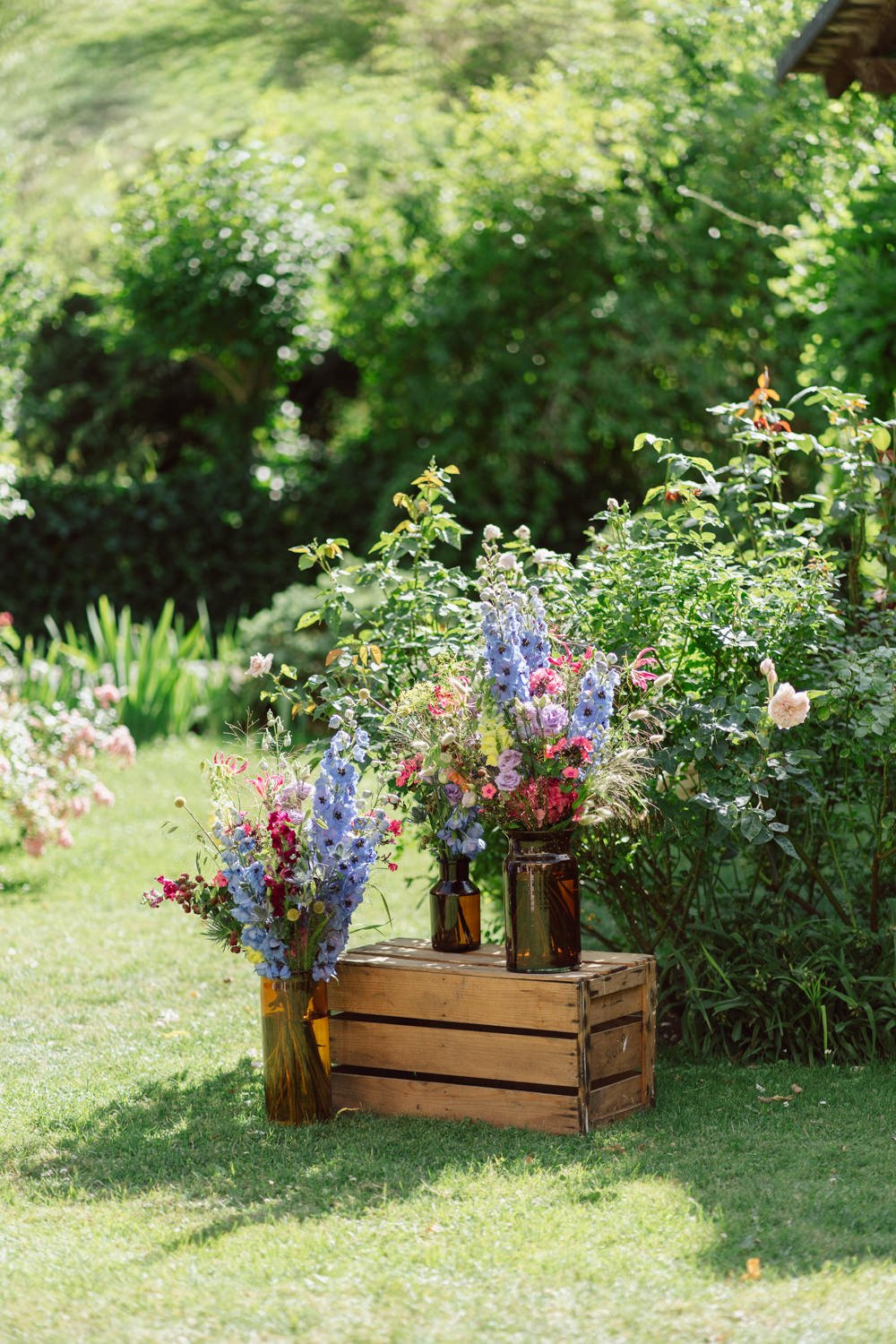 Fleurs colorées dans des vases en verre sur une caisse en bois, posées sur un gazon dans un jardin ensoleillé.