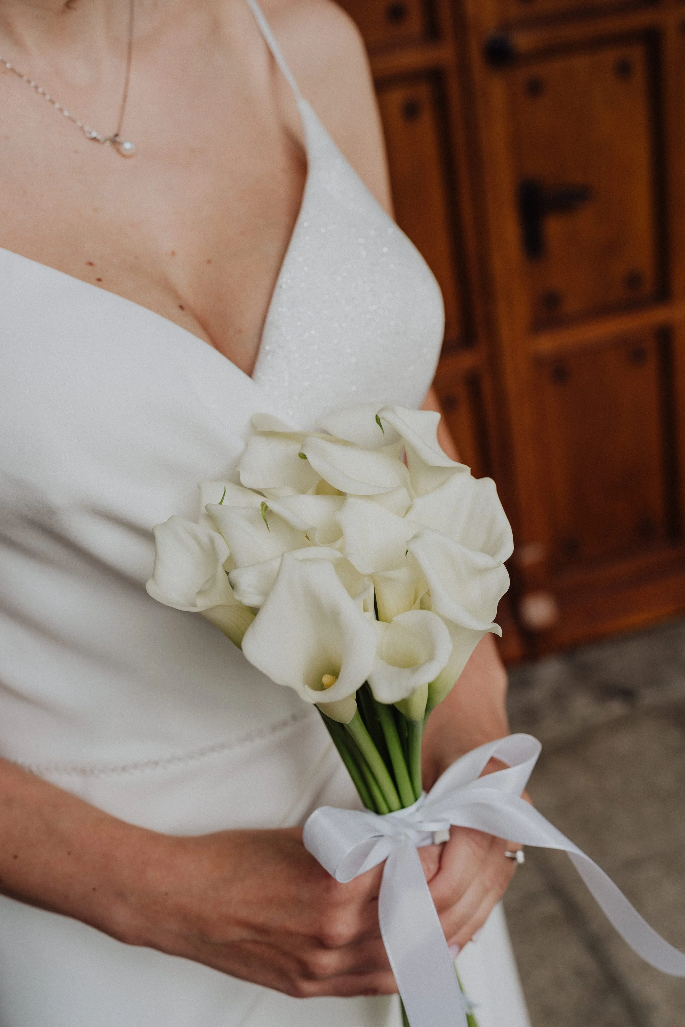 Un bouquet élégant de callas blanches tenu par une mariée, mettant en valeur la simplicité et la pureté de ce moment. La robe de mariée en satin et détails scintillants ajoute une touche de raffinement, tandis que l’arrière-plan en bois ancien crée u