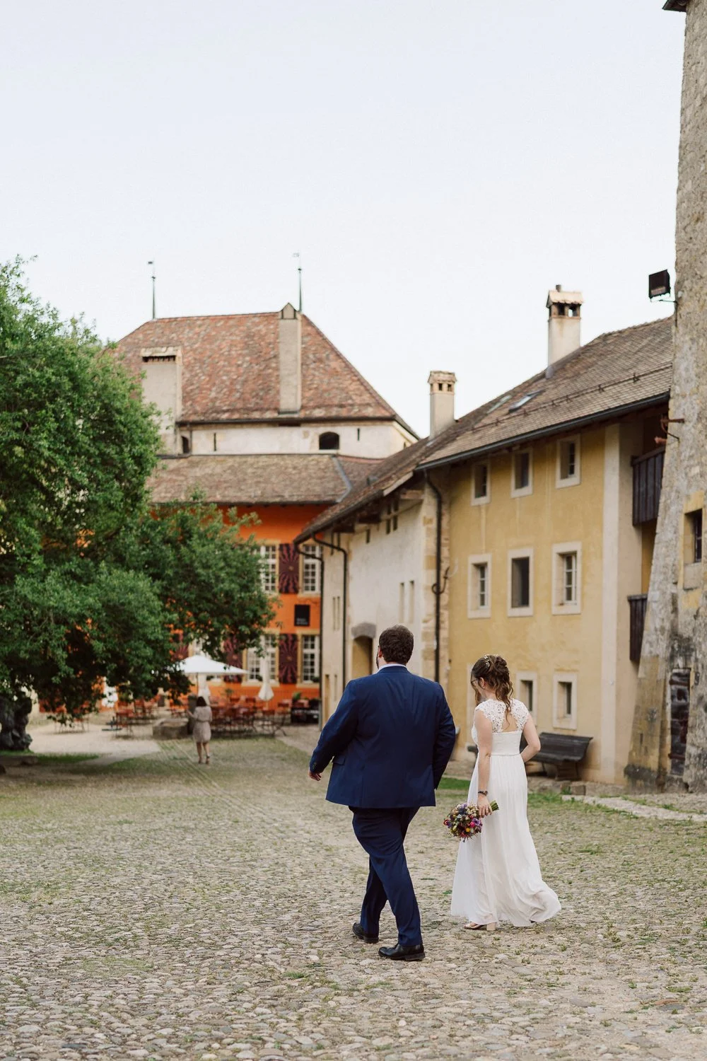 Couple de mariés marchant dans la cour du Jardin Baillival à Romainmôtier, robe de mariée fluide et bouquet coloré, cadre historique et ambiance intime, photo par Nonante Émotions.